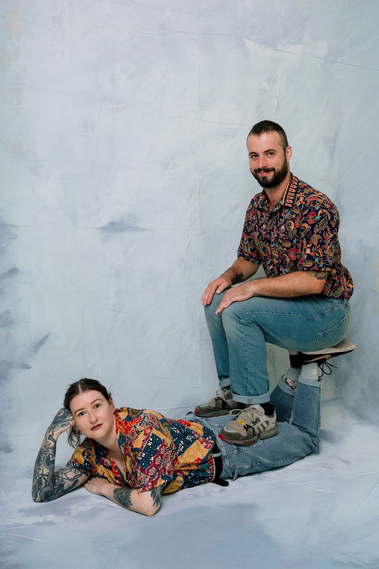 Two people in colorful floral shirts pose casually against a light gray backdrop in a studio setting.