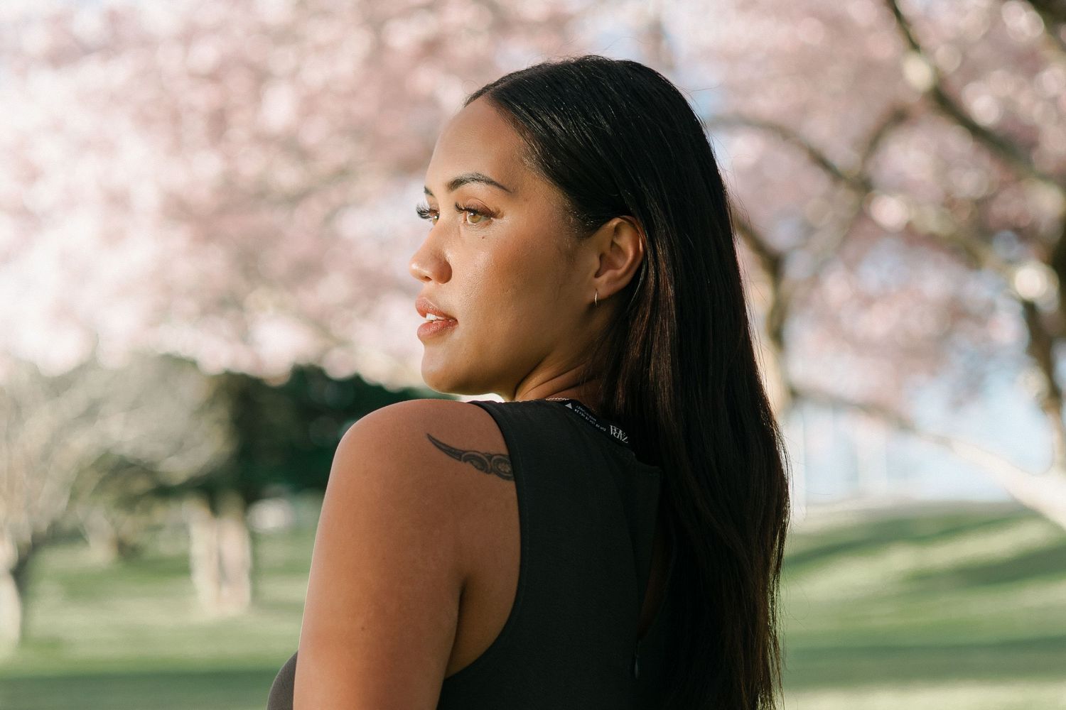 Woman with long dark hair poses in front of blooming cherry blossom trees during spring.