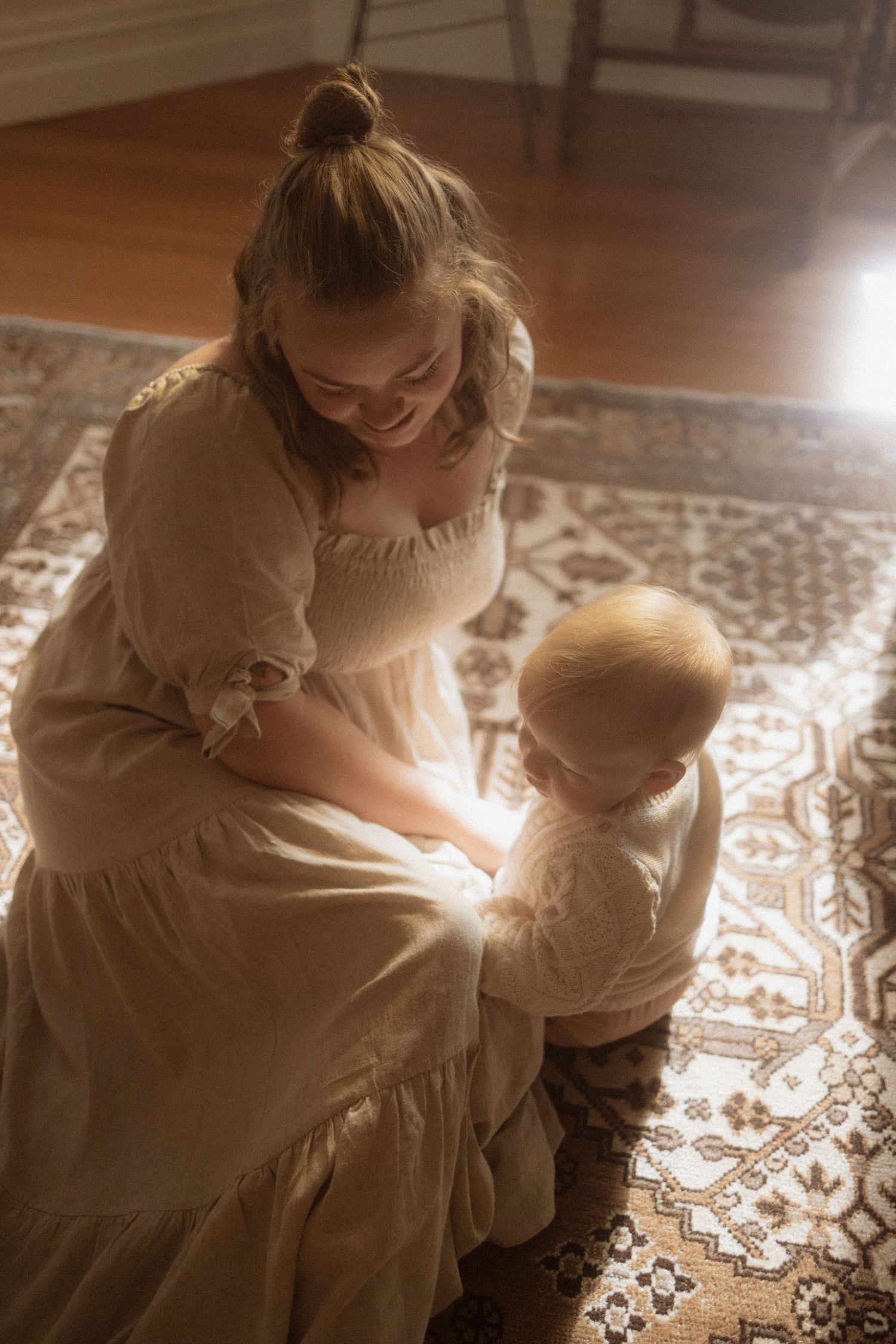 A tender moment captured between parent and child in vintage-style dresses on a patterned rug in warm lighting.