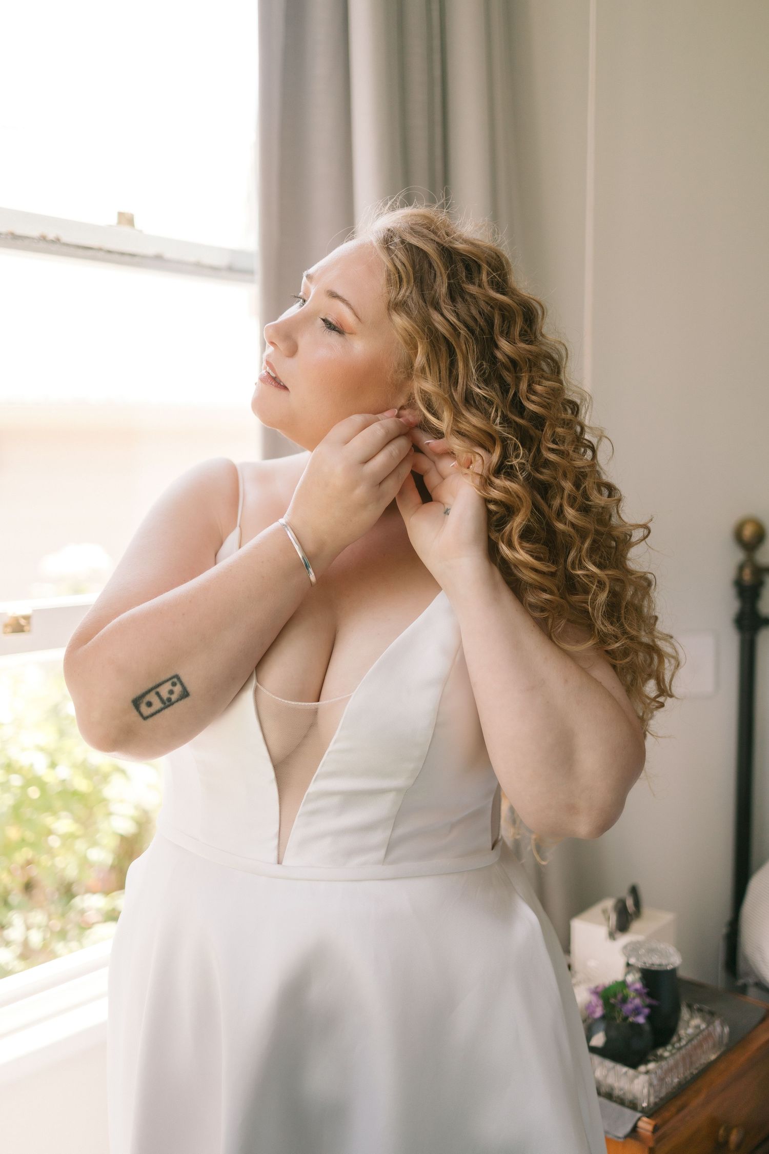 Person in white sleeveless top with curly hair fixing jewelry near window in soft natural light.