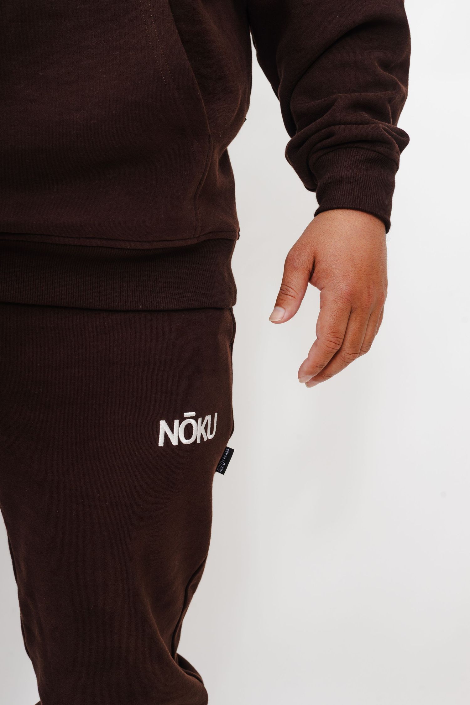 Close-up detail of brown sweatpants with white 'Now' text logo and matching brown sweatshirt sleeve visible.