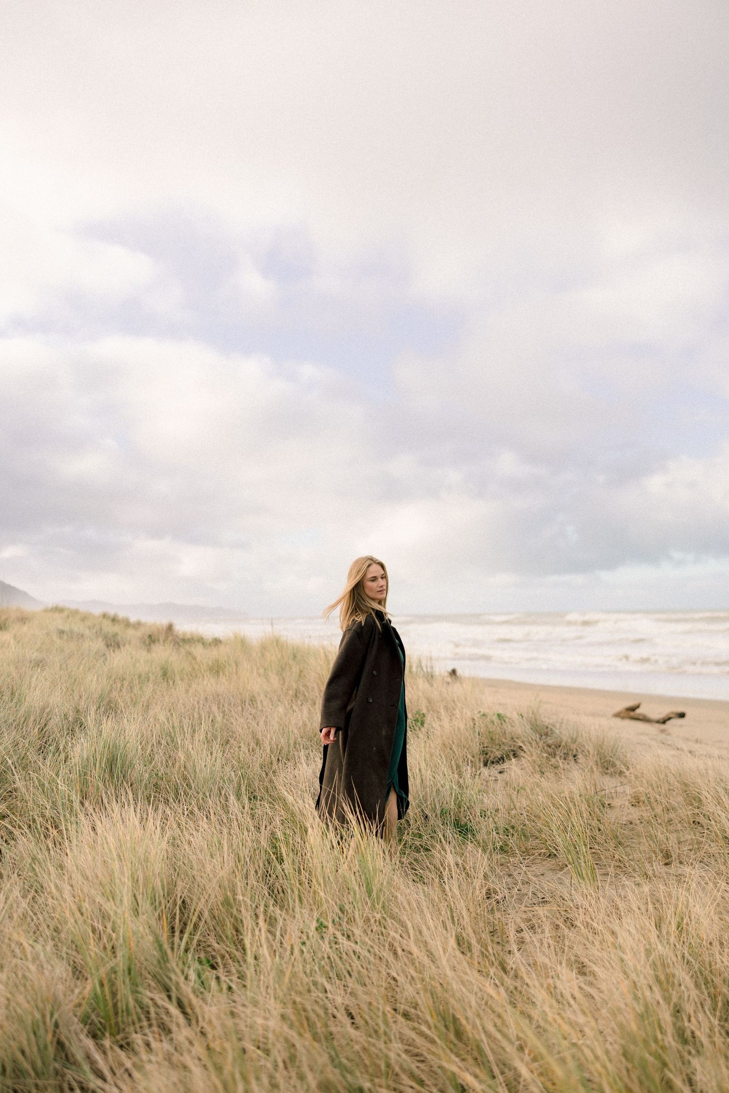 Woman in black coat standing among tall beach grasses with ocean waves and cloudy sky in background.