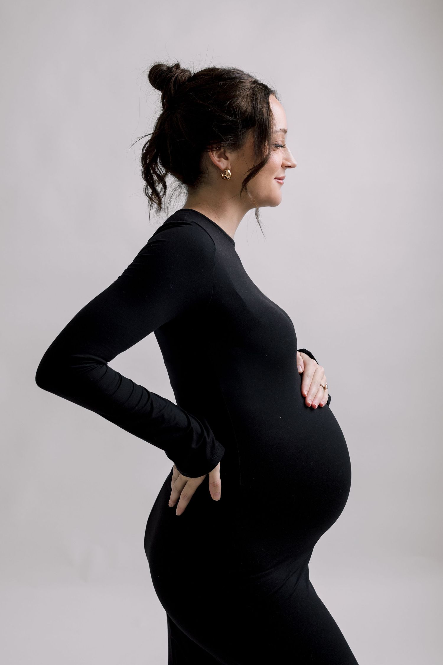 Smiling pregnant woman in black long-sleeve dress cradling baby bump, side profile view on gray background.