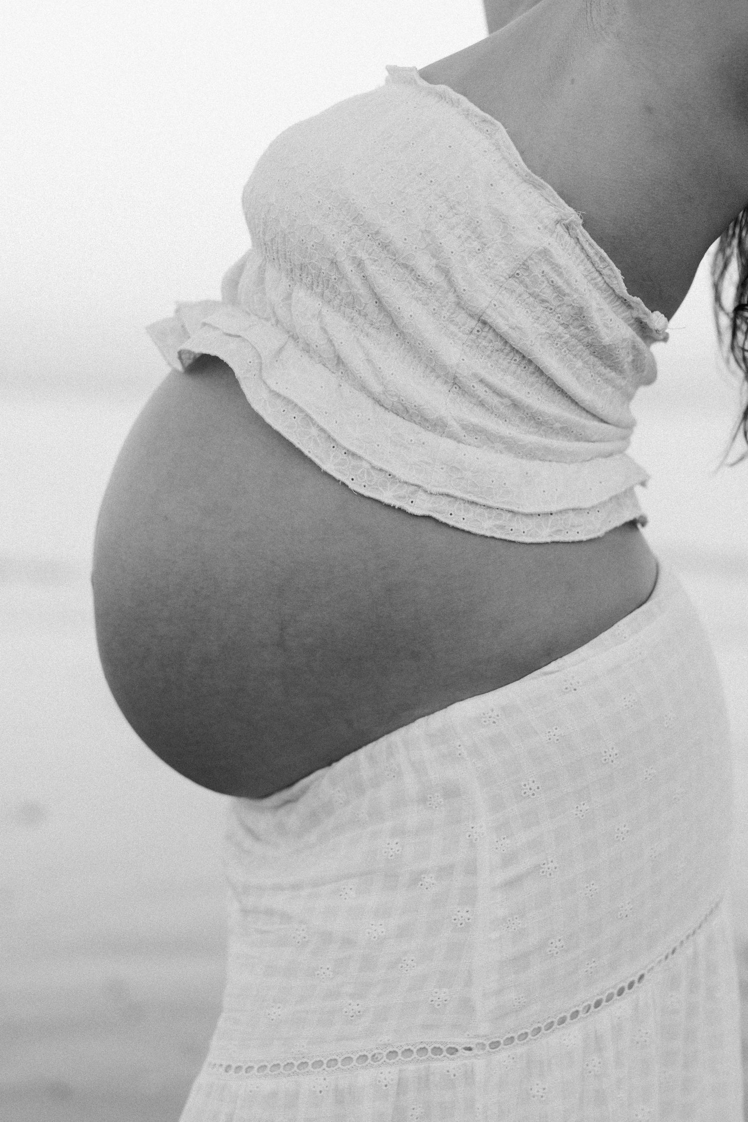 Black and white photo of a pregnant woman's bare baby bump in a white strapless top and skirt at the beach.