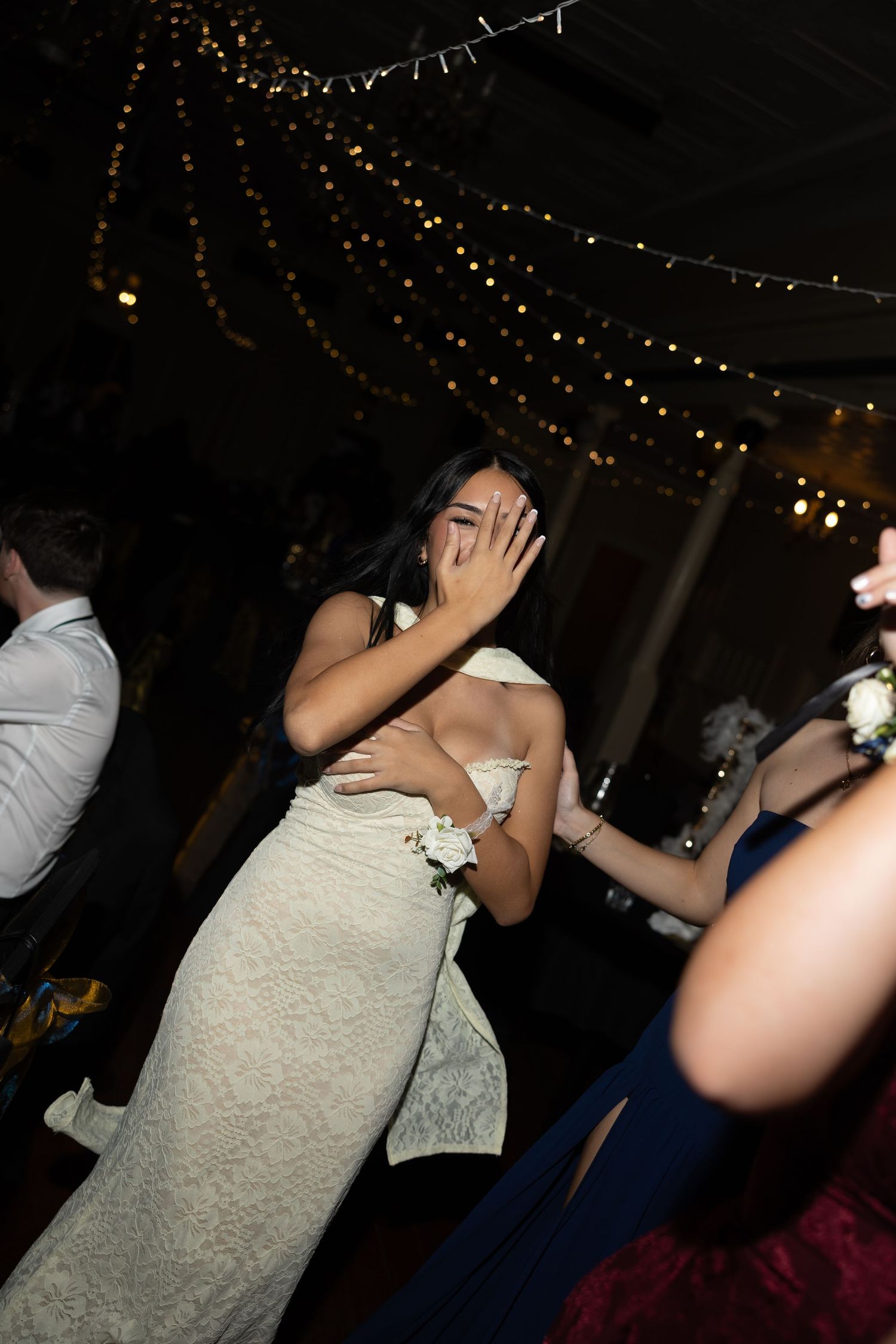 Woman in white off-shoulder gown covering her face while dancing at an elegant event with string lights.
