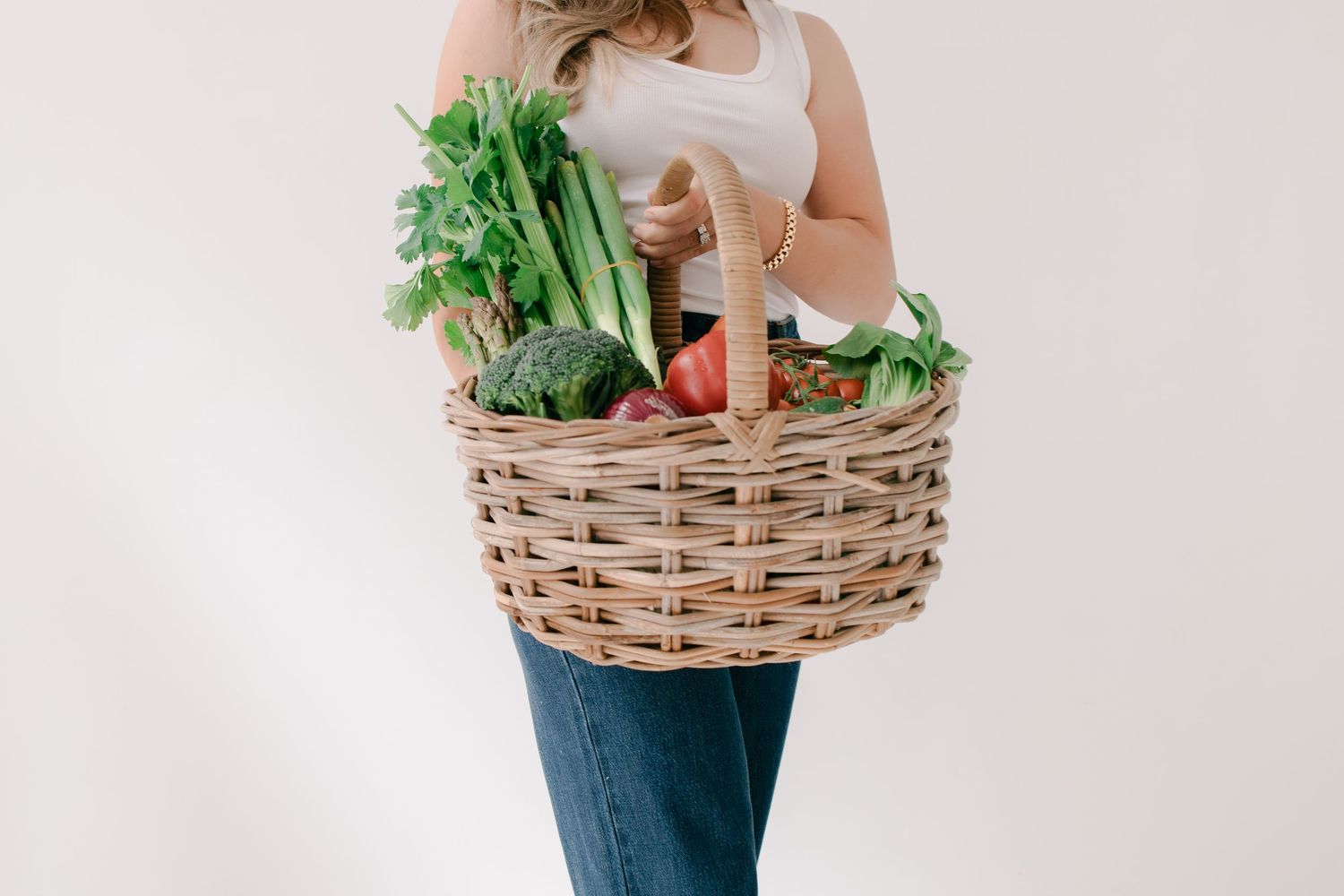 A woven basket filled with fresh vegetables and produce is held against a white background.