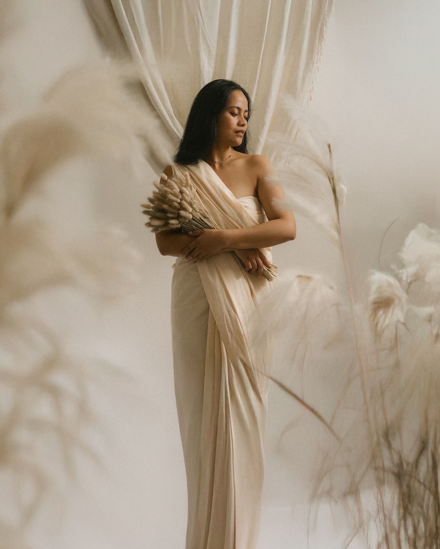 A graceful figure in a flowing cream gown holds dried pampas grass while posing against soft white drapes in a dreamy setting.