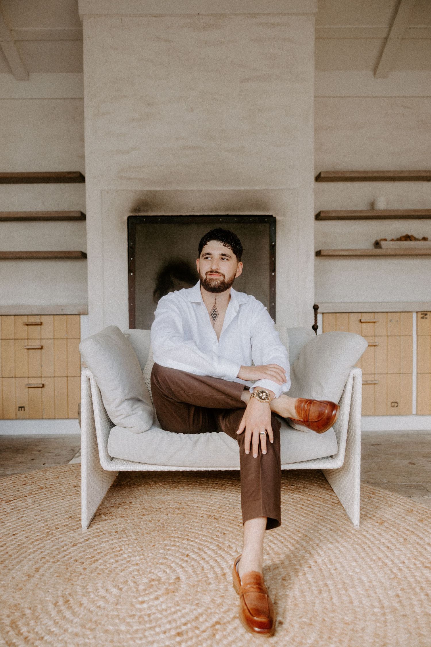 A person in a white shirt and brown pants sits relaxed in a modern white armchair on a circular jute rug.