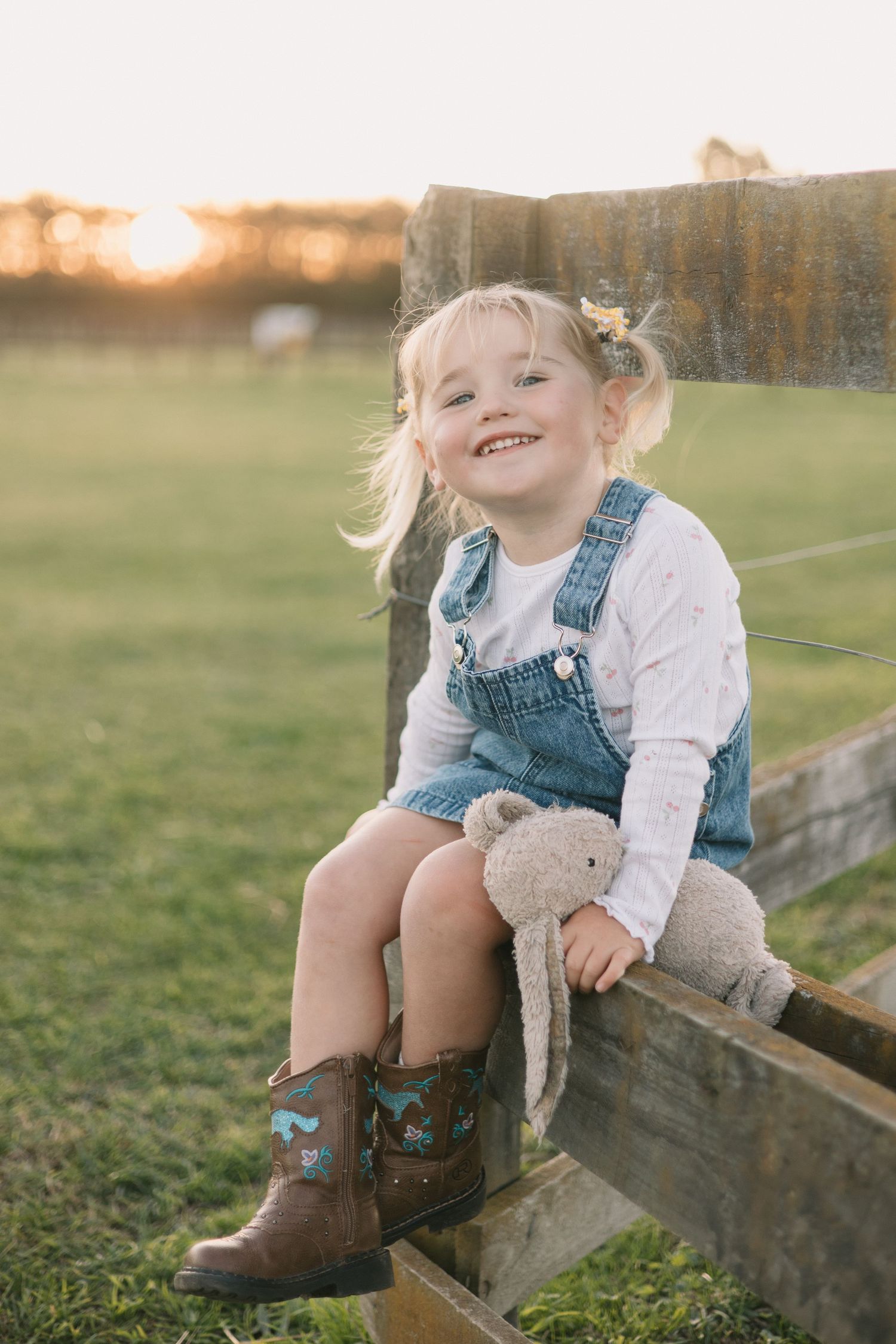 A child in denim overalls and cowboy boots sits on a wooden fence during sunset, holding a stuffed animal.