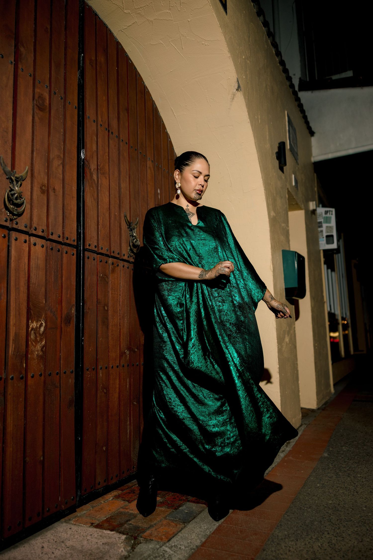 A person in an elegant emerald green velvet gown poses against a wooden door in a dimly lit hallway.