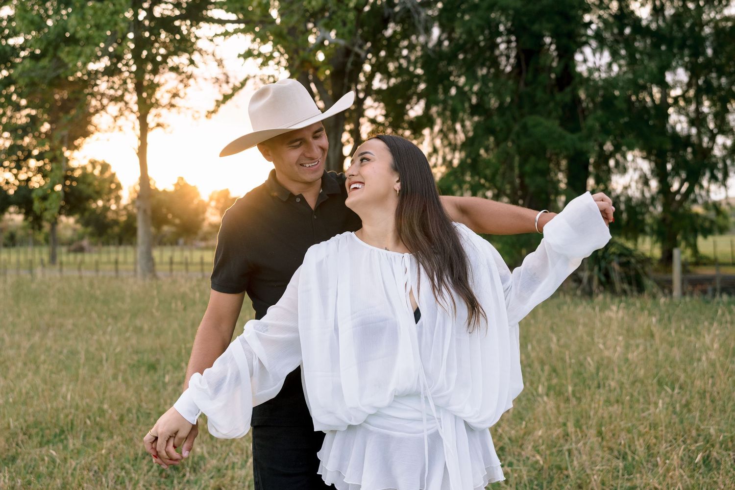 A couple embraces in a sunlit meadow at golden hour wearing white and black outfits.