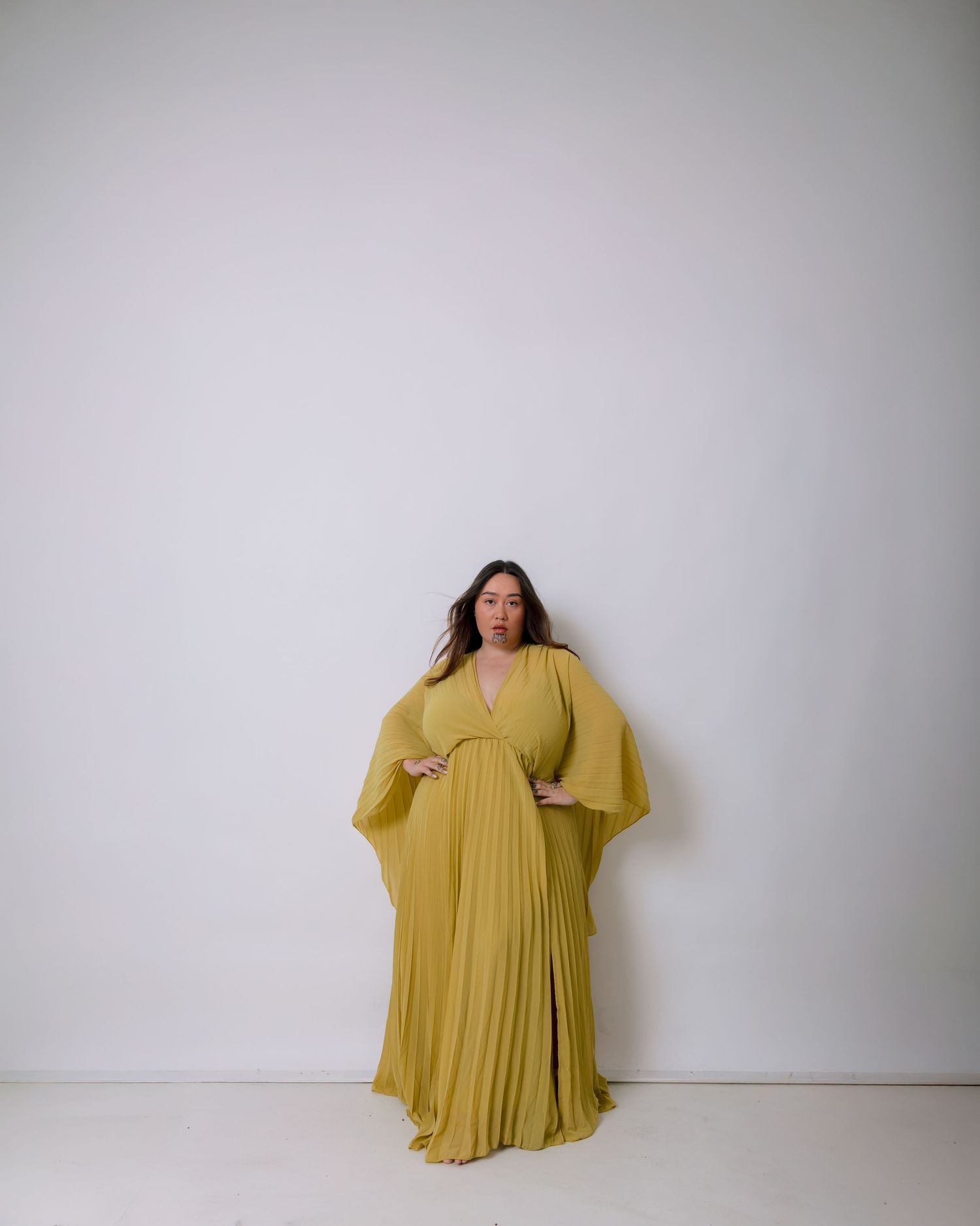 A model in a flowing mustard yellow dress poses against a white wall showing the garment from multiple angles.