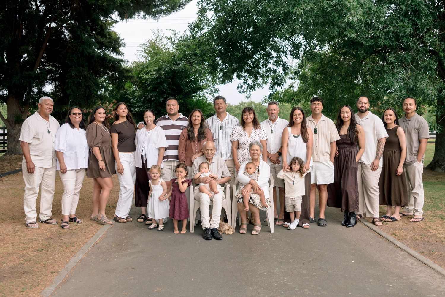 Large multigenerational family group portrait outdoors on a path surrounded by lush green trees.
