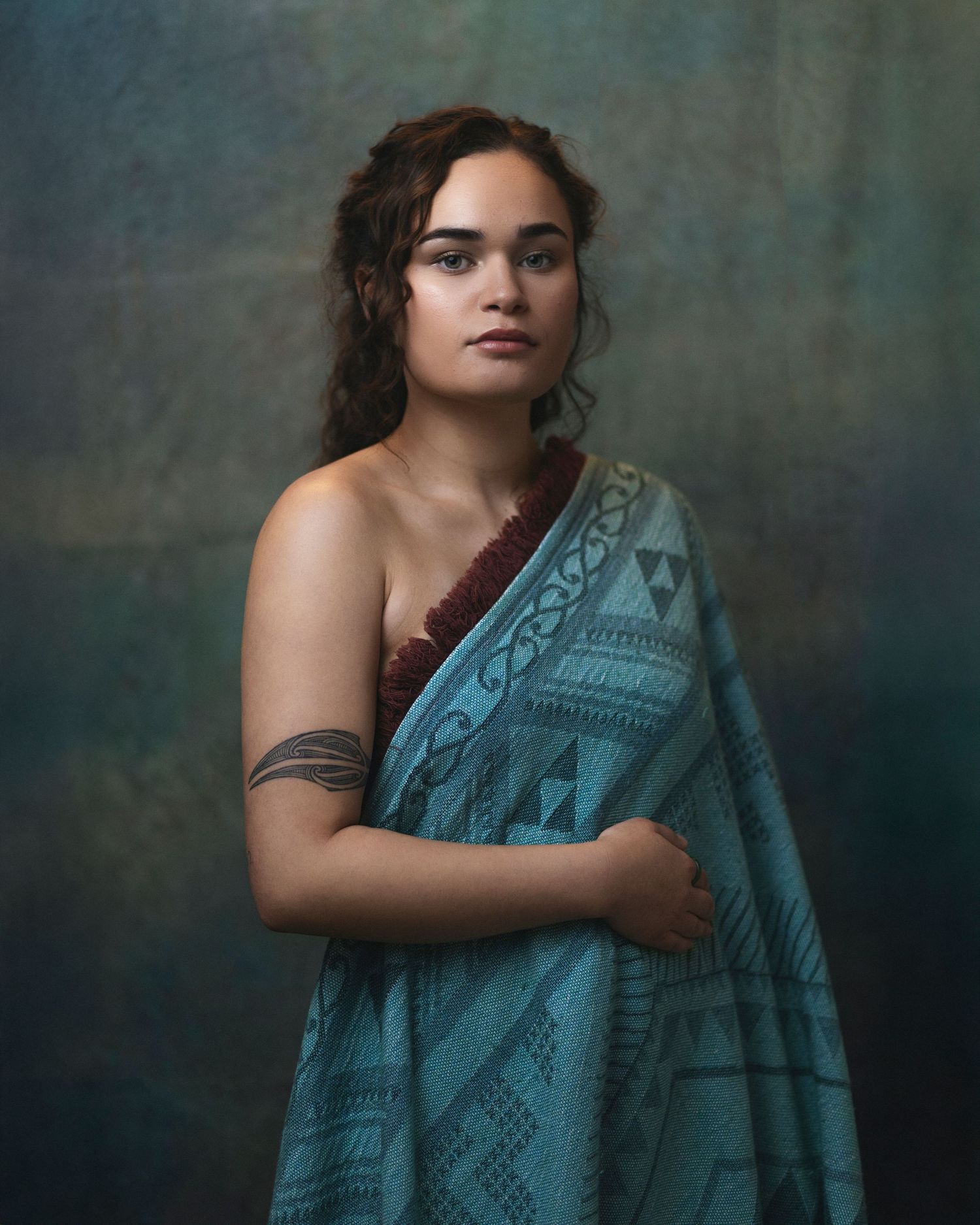 Woman in teal patterned wrap with tribal arm tattoo and red necklace poses against moody blue-gray backdrop.
