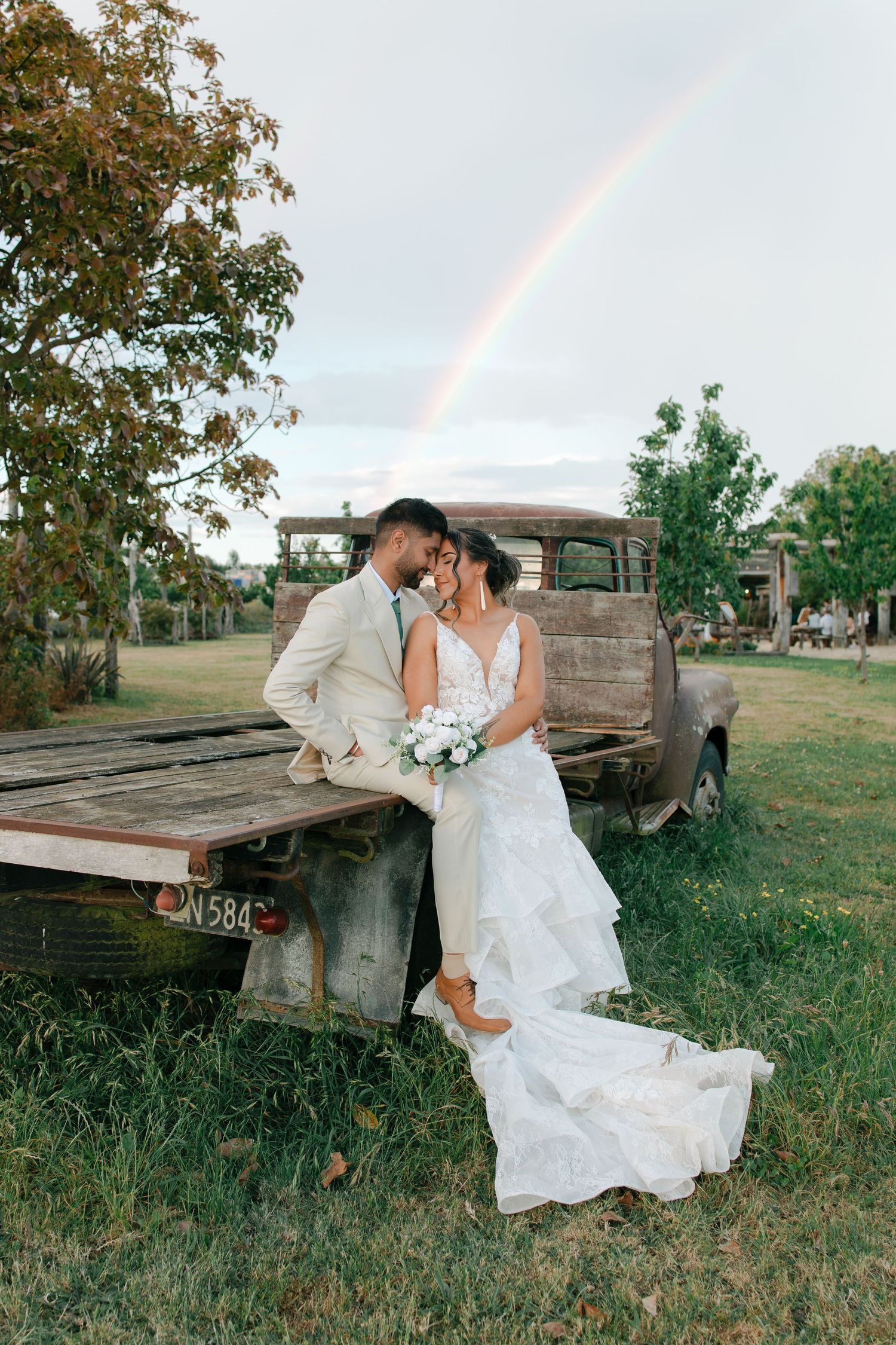 A romantic couple in wedding attire pose together near a rustic wooden wagon in a grassy field at sunset.