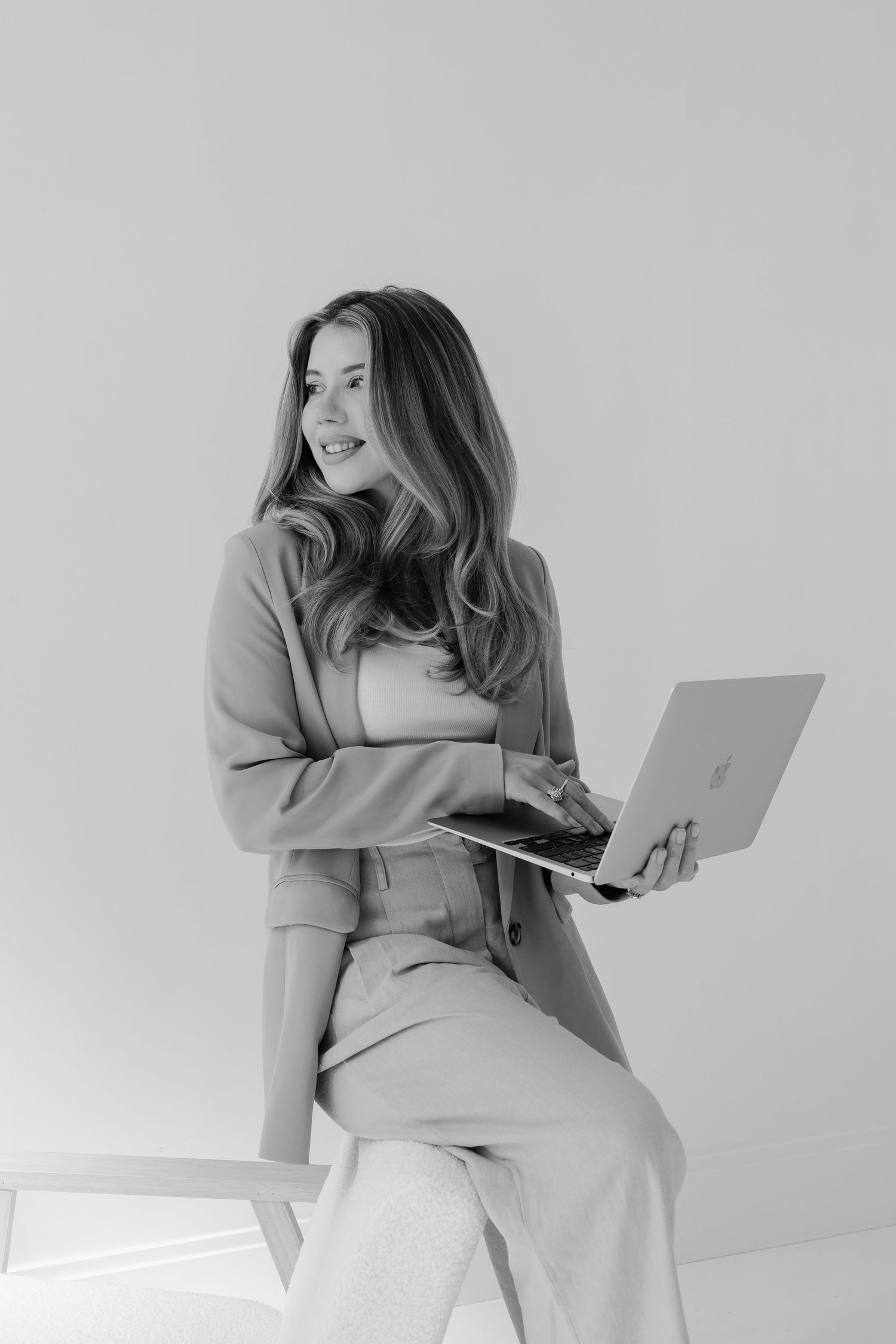 Black and white photo of someone in casual business attire sitting with a laptop and smiling against a plain background.