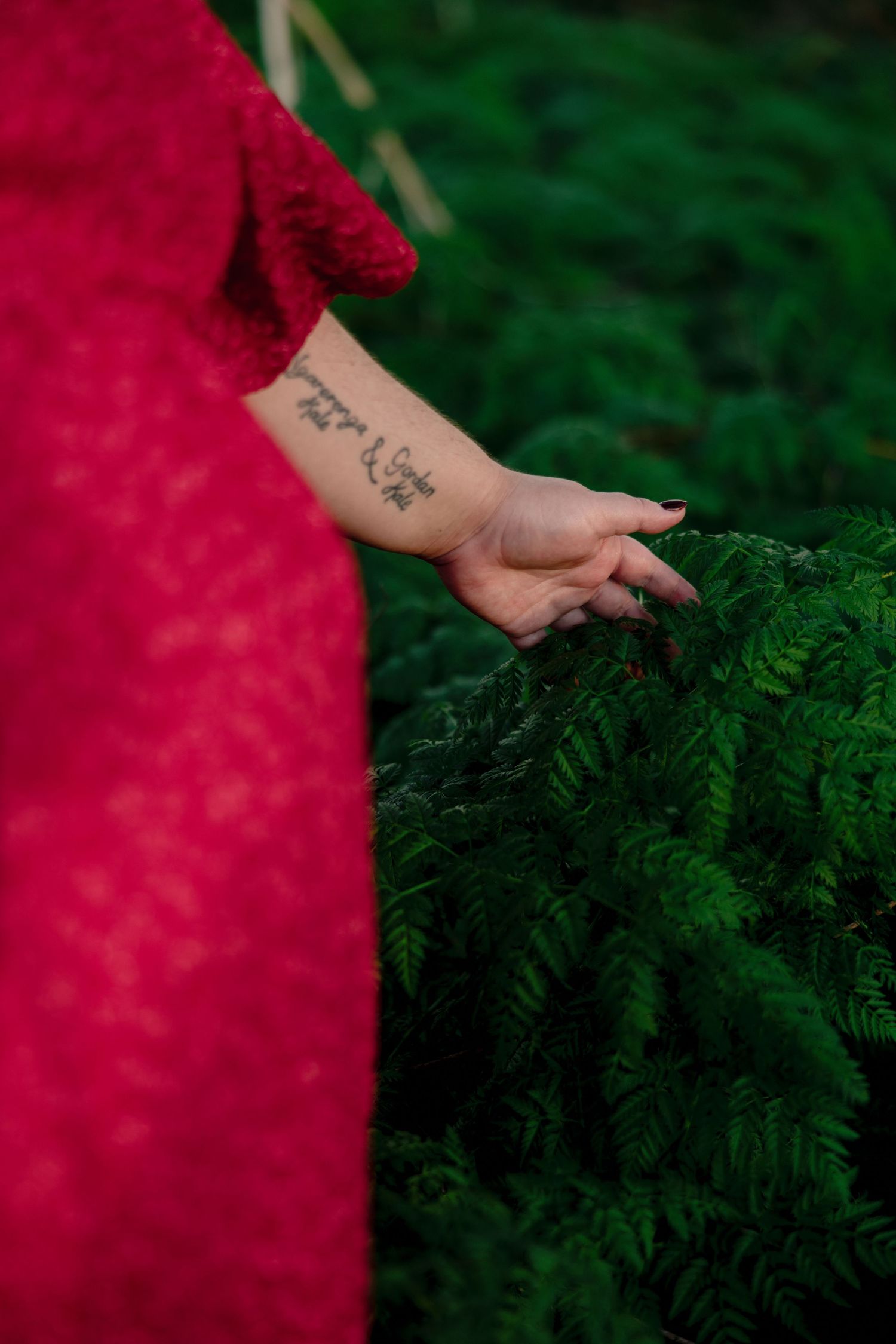 Woman in red lace dress touching green ferns with tattooed forearm in a lush garden setting.