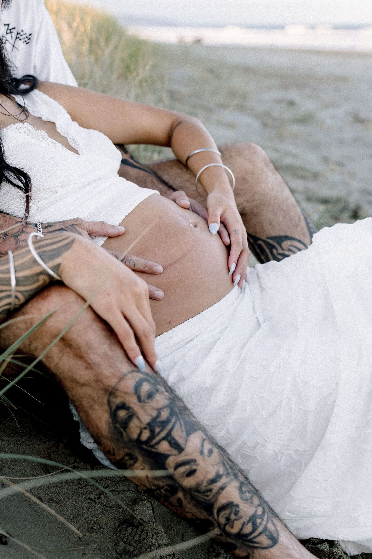 Tattooed couple sitting on beach sand, hands tenderly cradling pregnant belly, wearing white outfits.