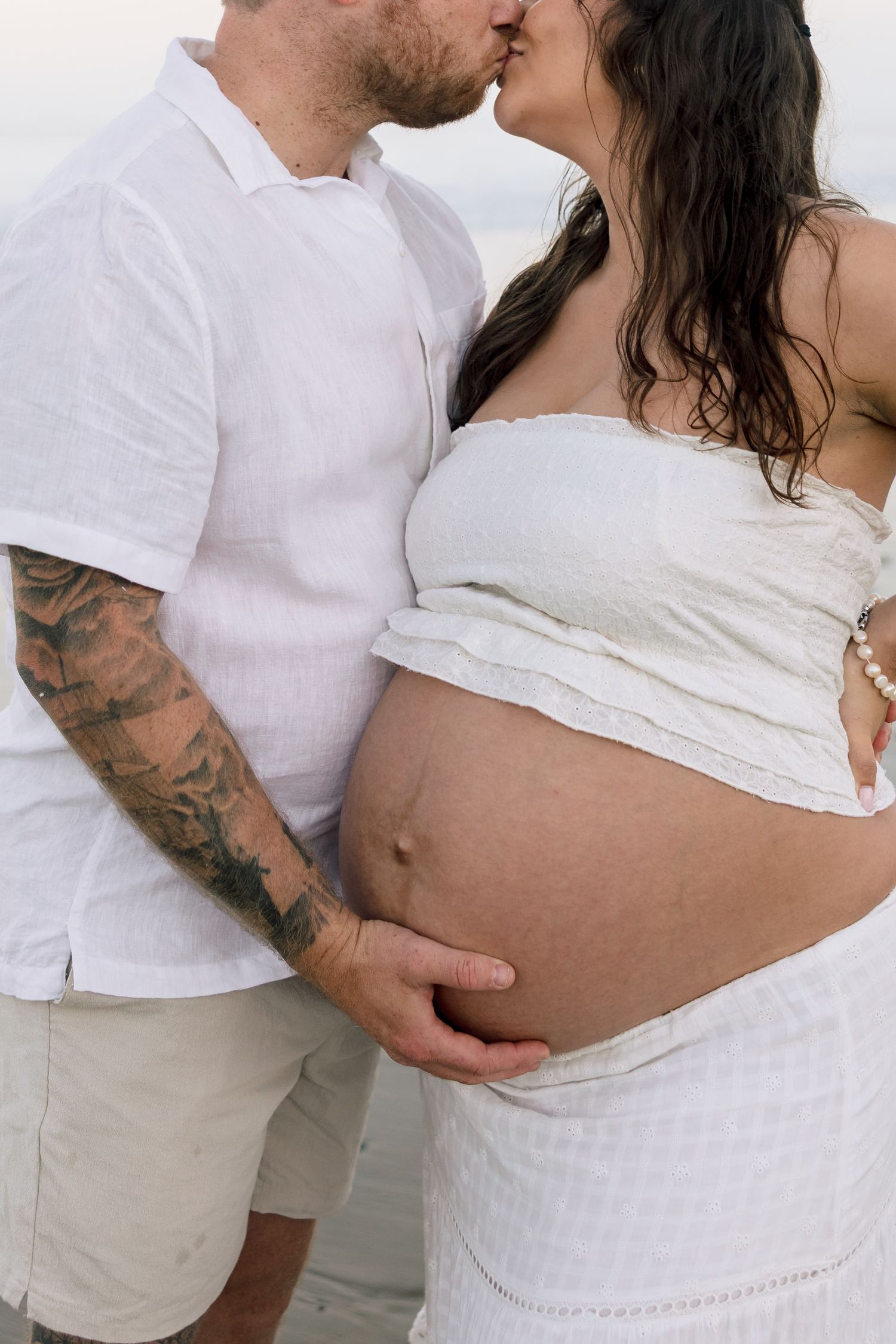 Couple in white outfits sharing a kiss while cradling pregnant belly on a beach.