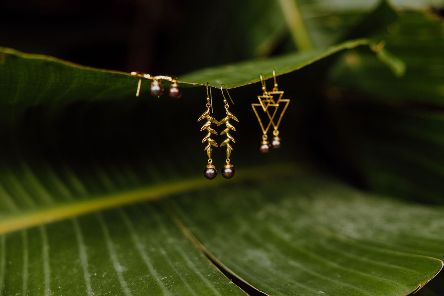 Delicate gold tribal-style earrings displayed hanging on a leaf against a dark green blurred background.