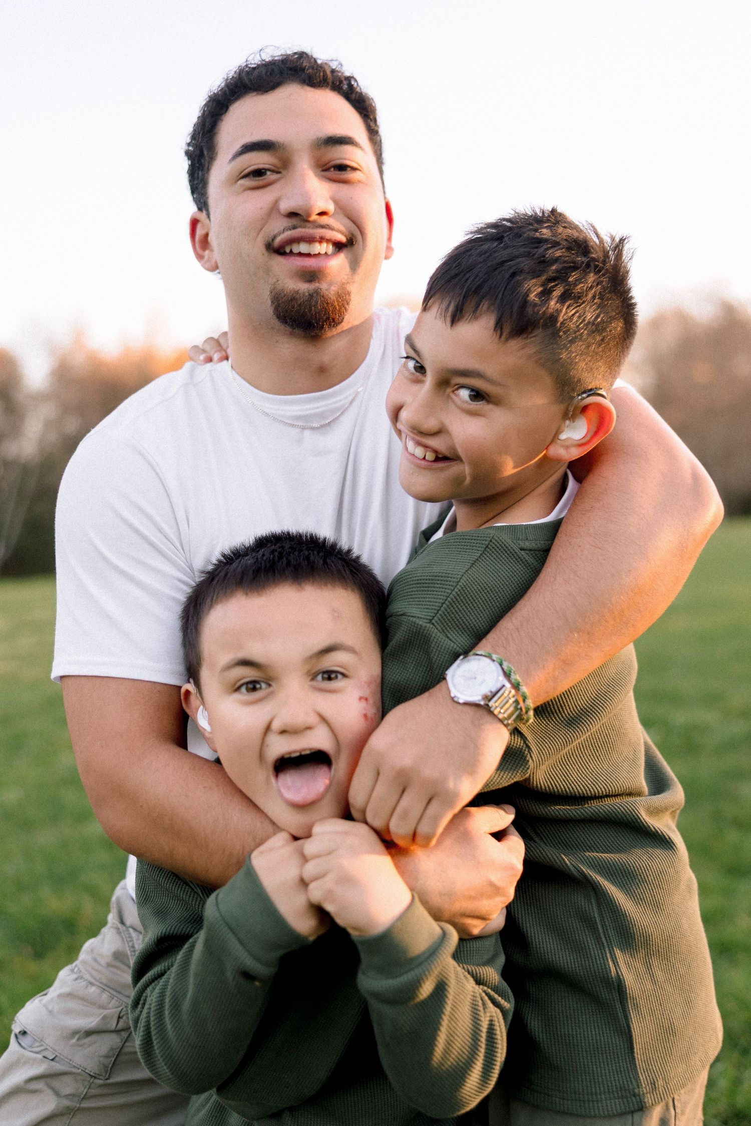 A young man smiles while playfully hugging two boys outdoors in a sunny park setting.