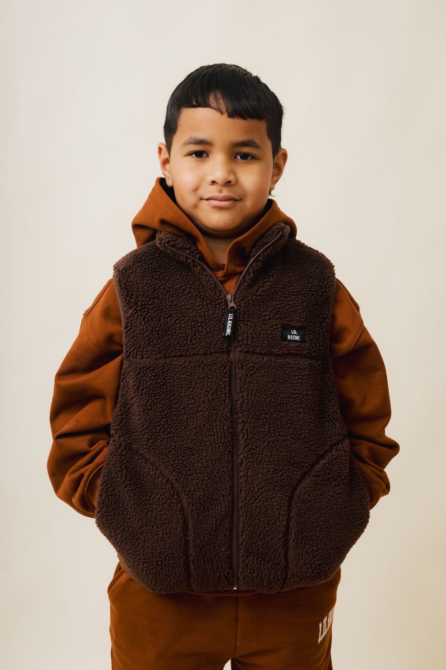 Child wearing brown fuzzy fleece vest with light brown hoodie sweatshirt against neutral background.