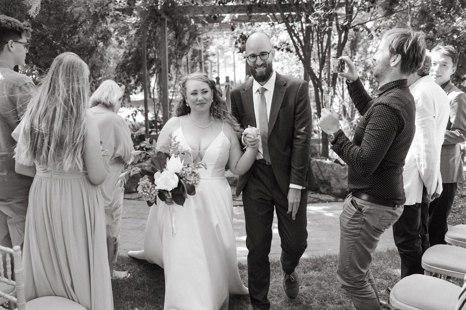 Black and white photo showing guests at an outdoor garden wedding ceremony with a couple and attendants gathered in celebration.