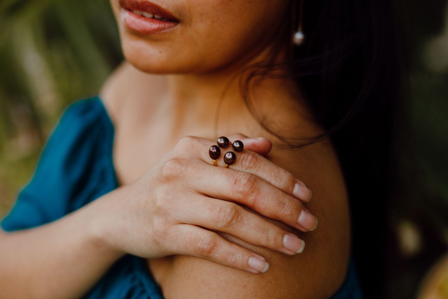 Close-up view of hands with natural manicure and dark ornate ring resting on shoulder in teal blue garment.