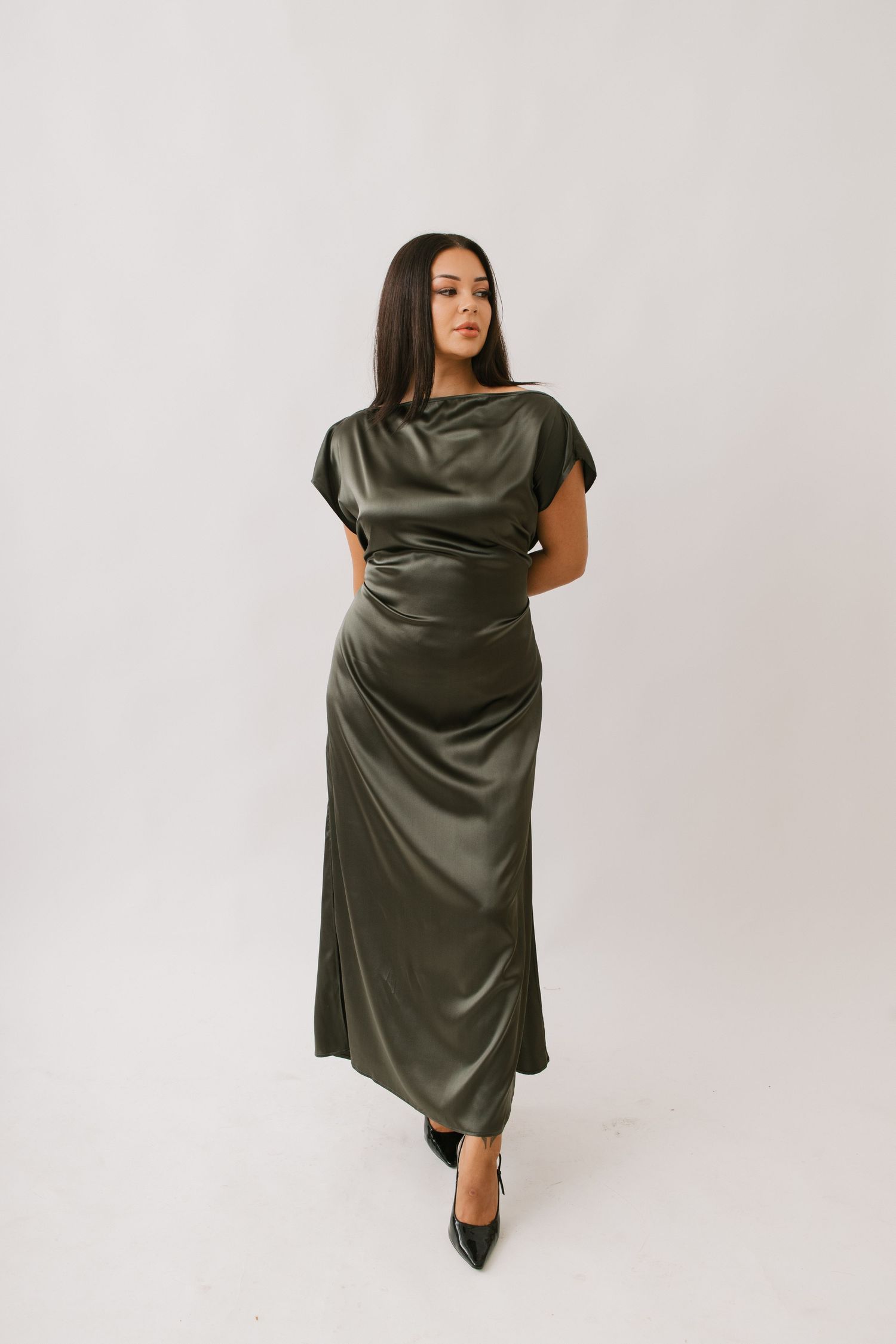 Woman poses in an elegant olive green satin dress with cap sleeves and ruched detailing against a white backdrop.