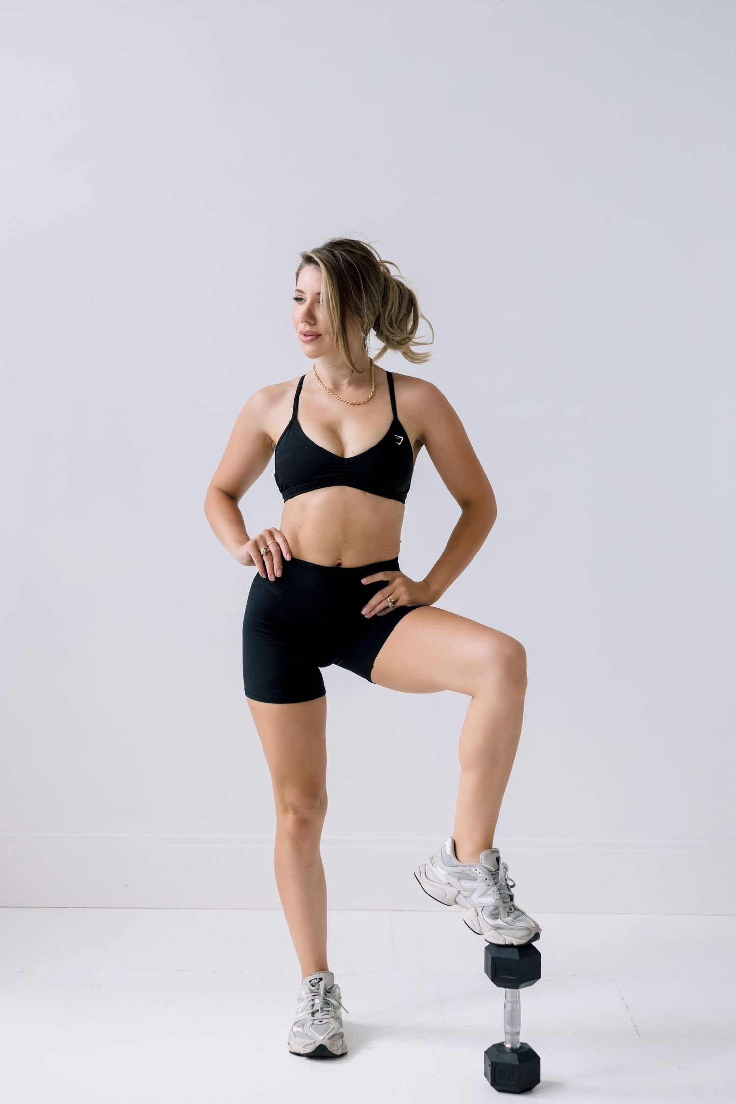 Woman in black sports bra demonstrating lunge exercises with dumbbell weight against light backdrop.