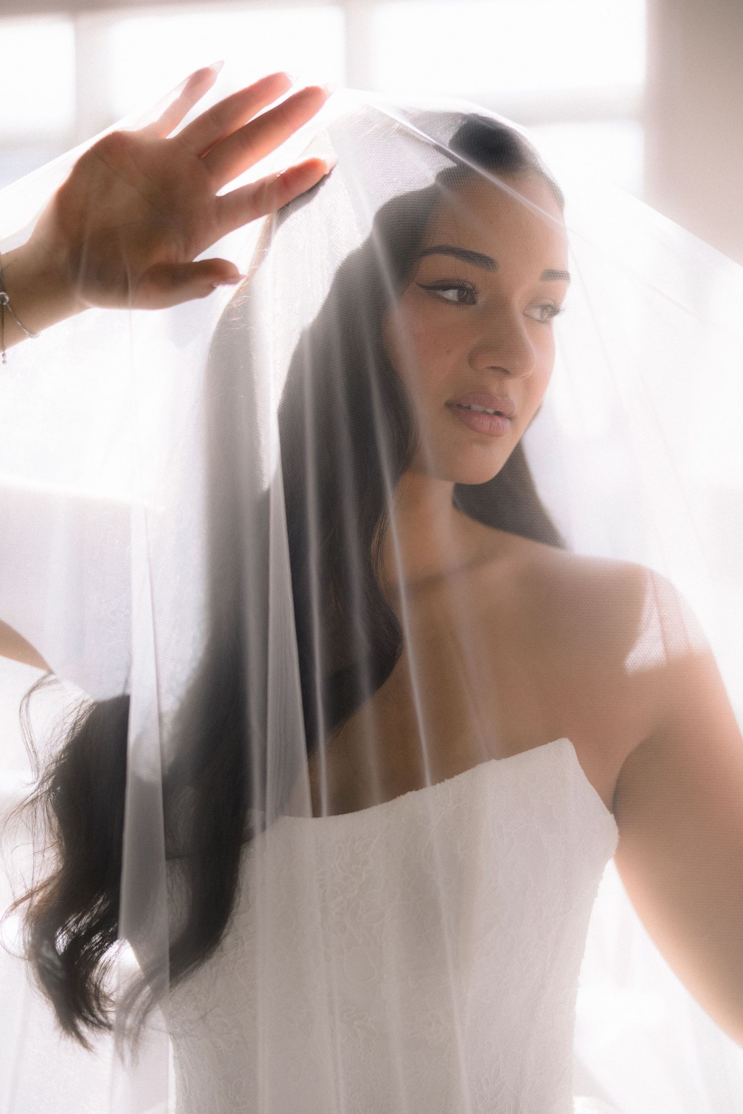 Bride in white strapless gown lifting sheer veil, soft backlit portrait with elegant bridal look.