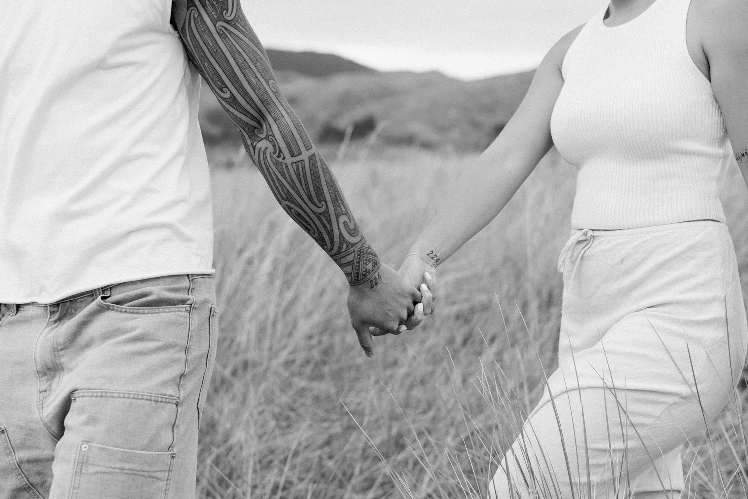 Two people in white outfits holding hands while walking through a grassy meadow on an overcast day.