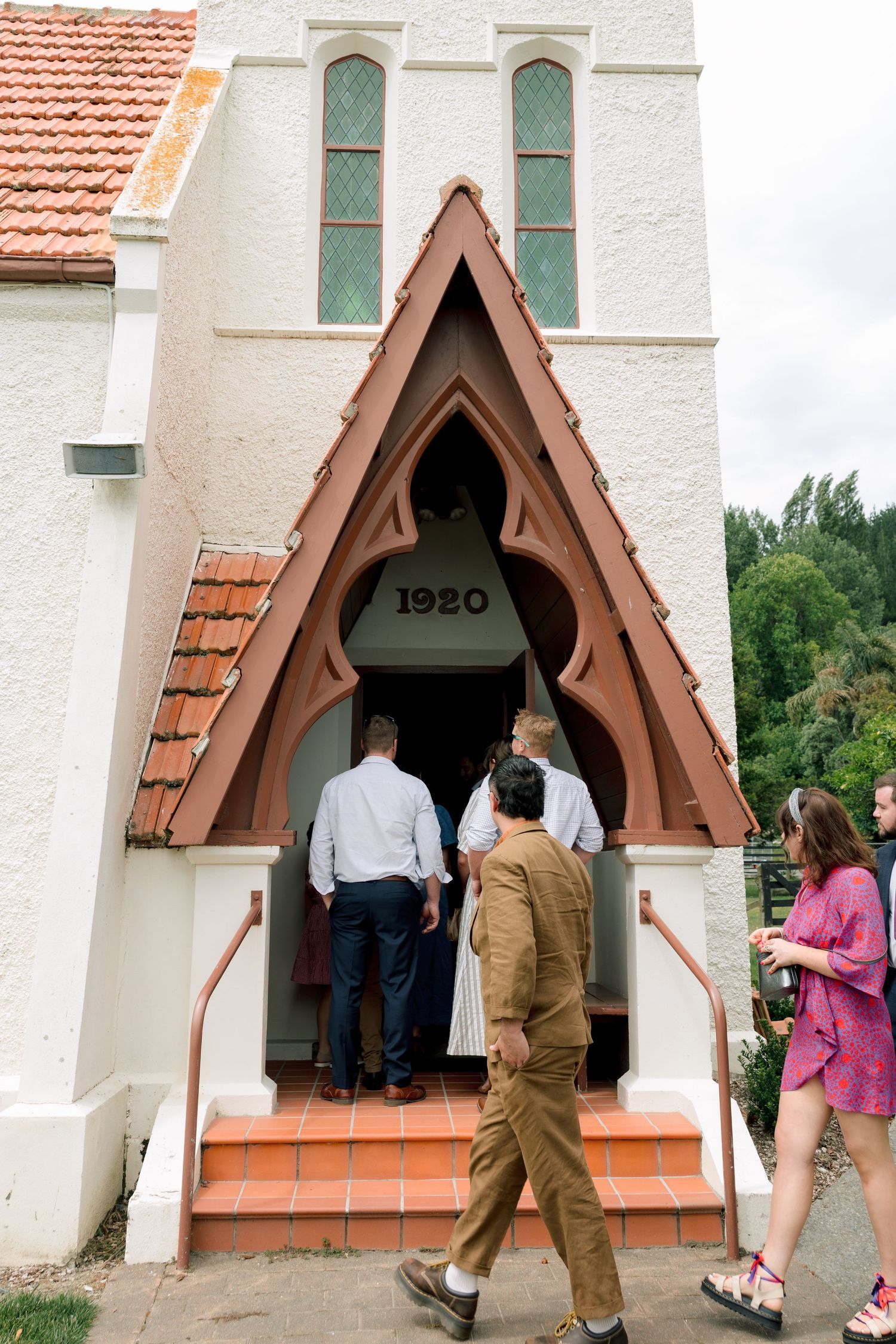 People entering a small white church with a triangular brown entrance and the year 1920 marked above the doorway.