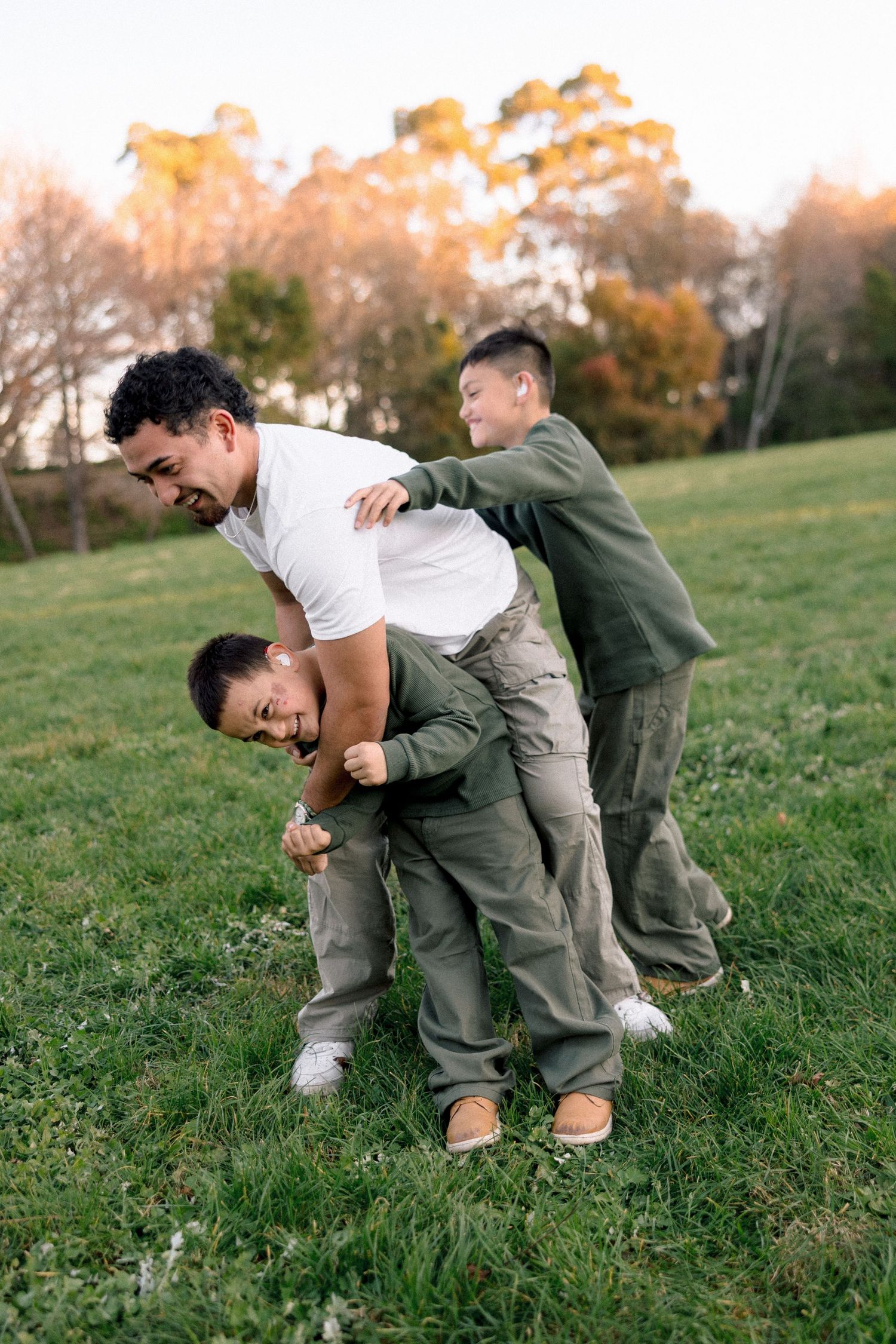 Father and two boys playing and laughing together on green grass in a park during golden hour.