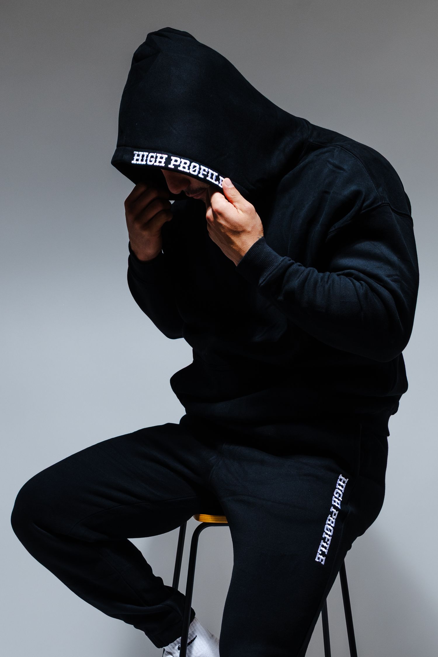 Model in black High Profile streetwear ensemble sits on stool covering face against studio backdrop.