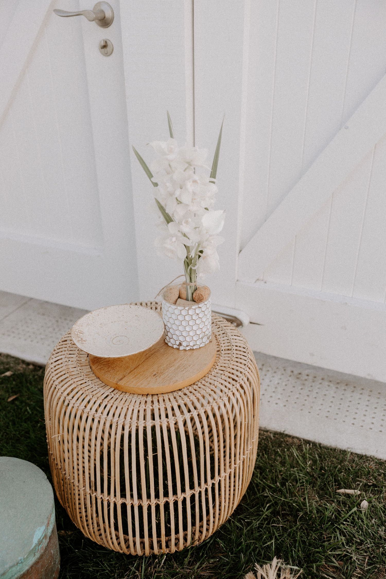 Rattan side table with white orchid arrangement in woven vase and decorative plate on wooden top against white door.