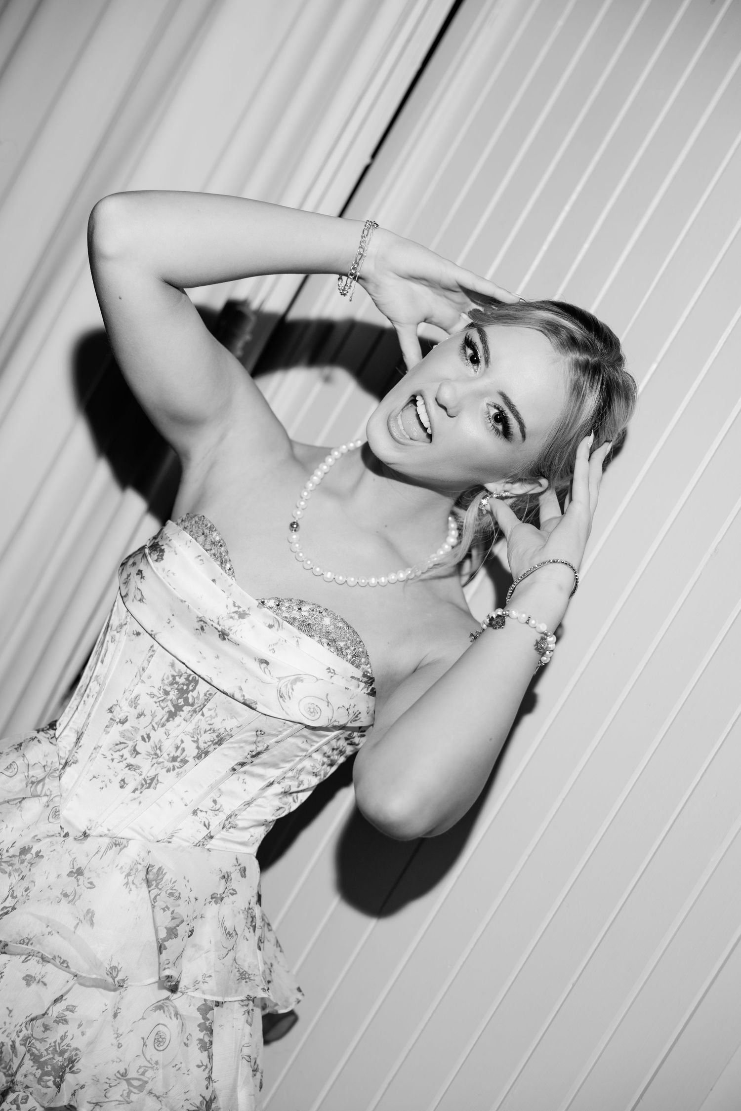 Elegant blonde woman in floral strapless dress with pearl necklace posing gracefully in black and white photo.