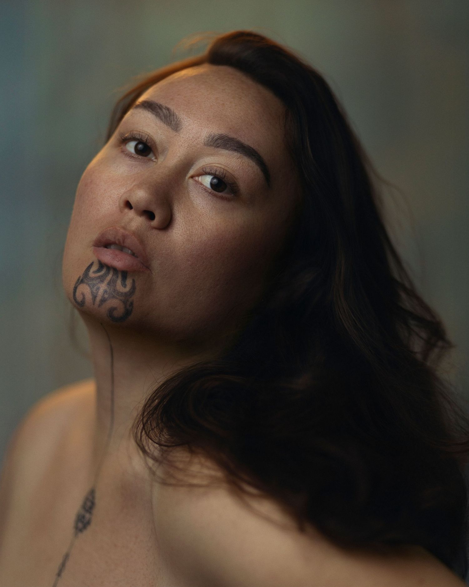 Portrait of a woman with traditional Maori chin tattoo and long dark hair against a muted background.