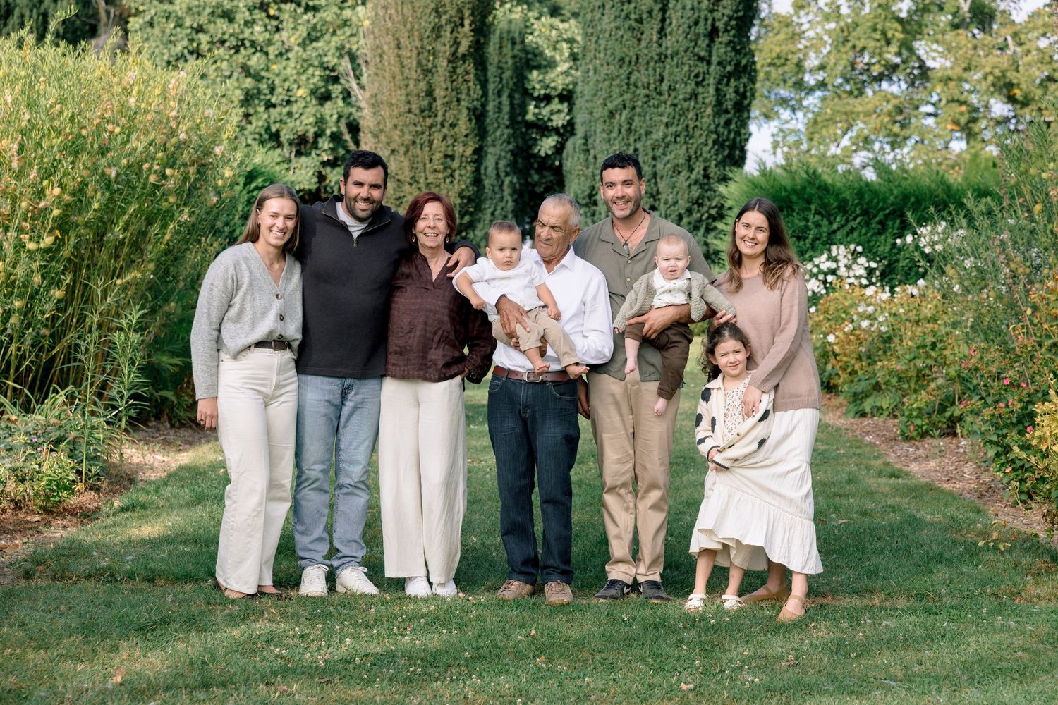 Multi-generational family of seven smiling together outdoors in a lush green garden, including two young children.
