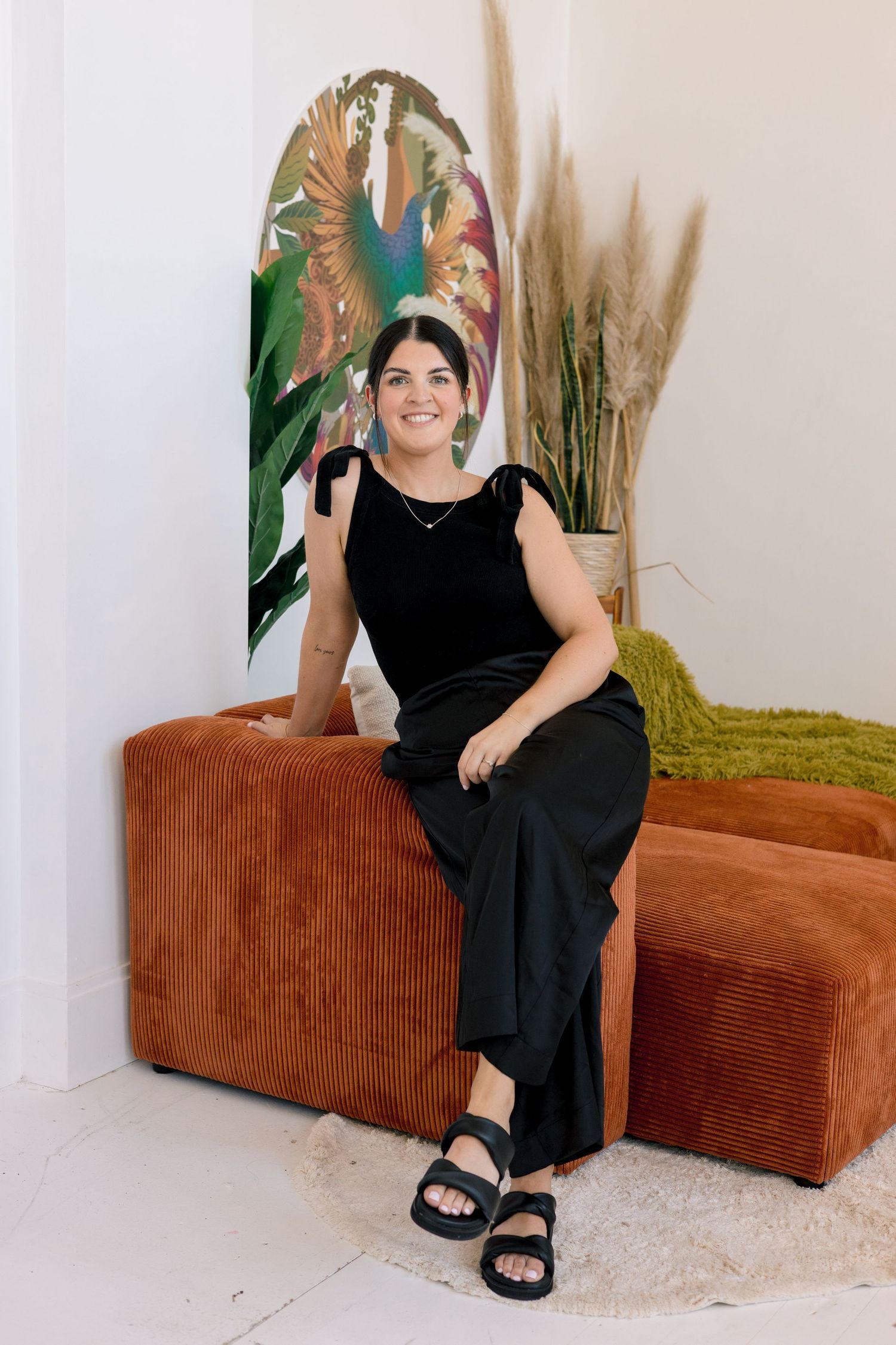 A person in a black dress sits on a brown corduroy couch with indoor plants in the background.