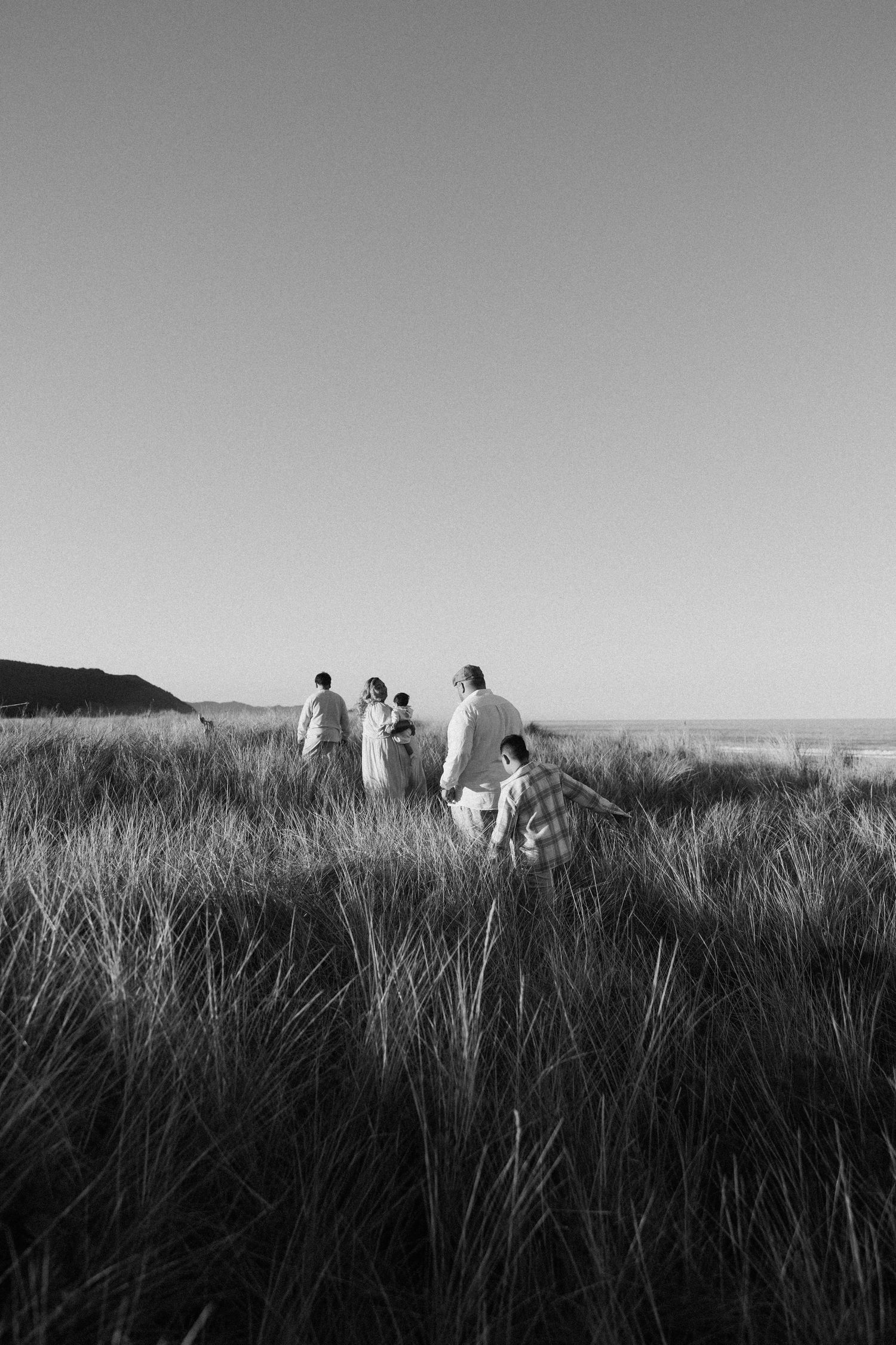 A family walks together through tall grass dunes in a black and white coastal landscape photo.