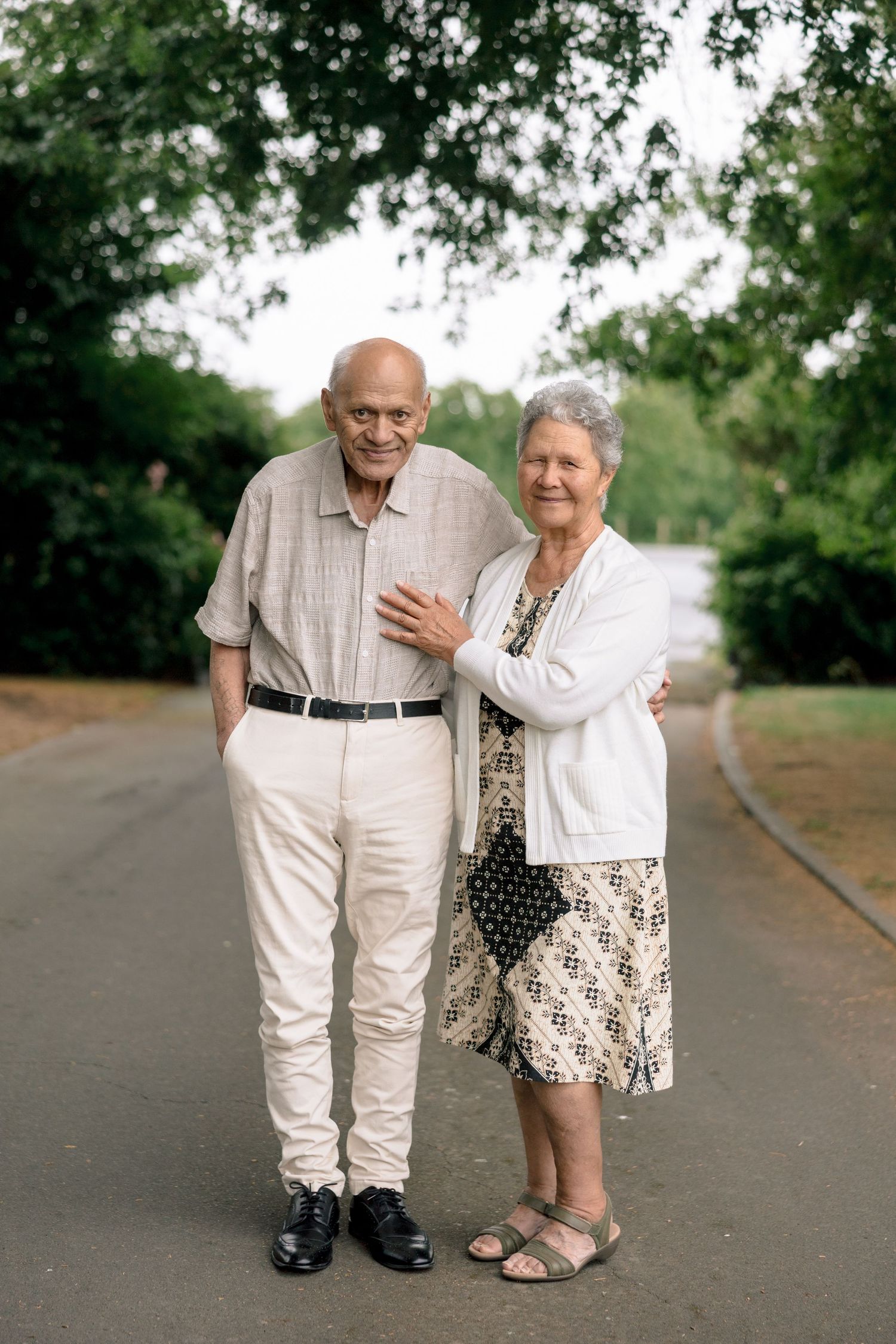 An elderly couple walks together on a path, wearing light colored clothing and holding each other tenderly.
