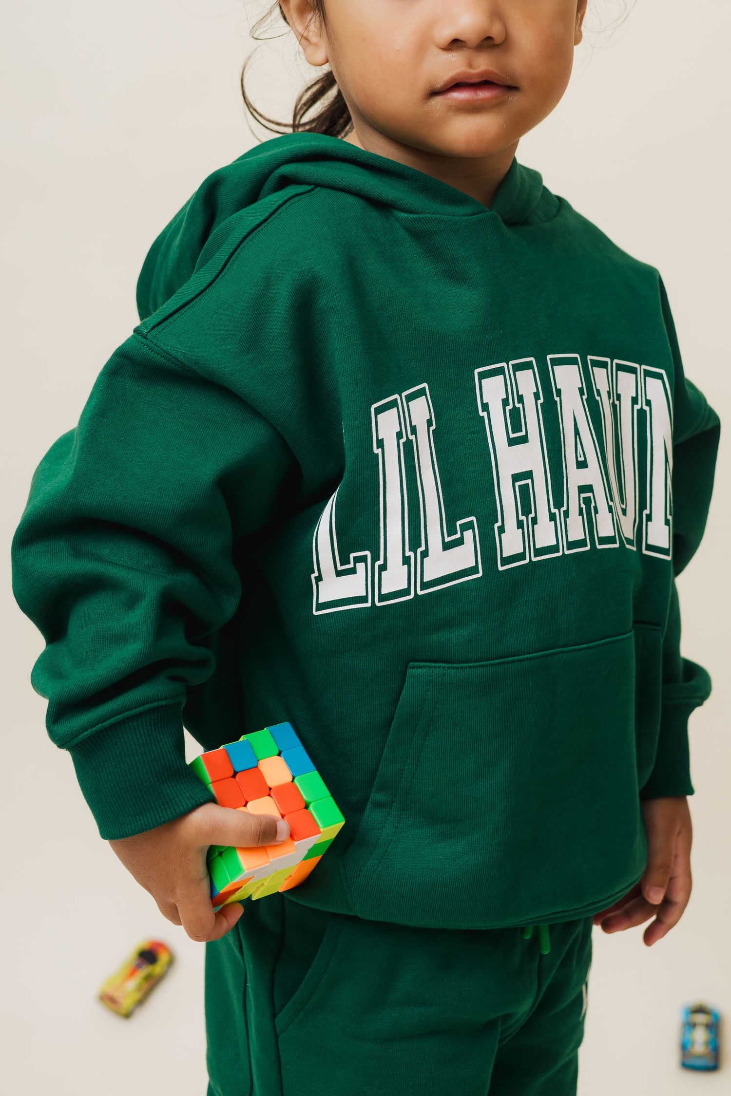 A small child wearing a green Harvard sweatshirt holds a colorful toy while standing against a light background.