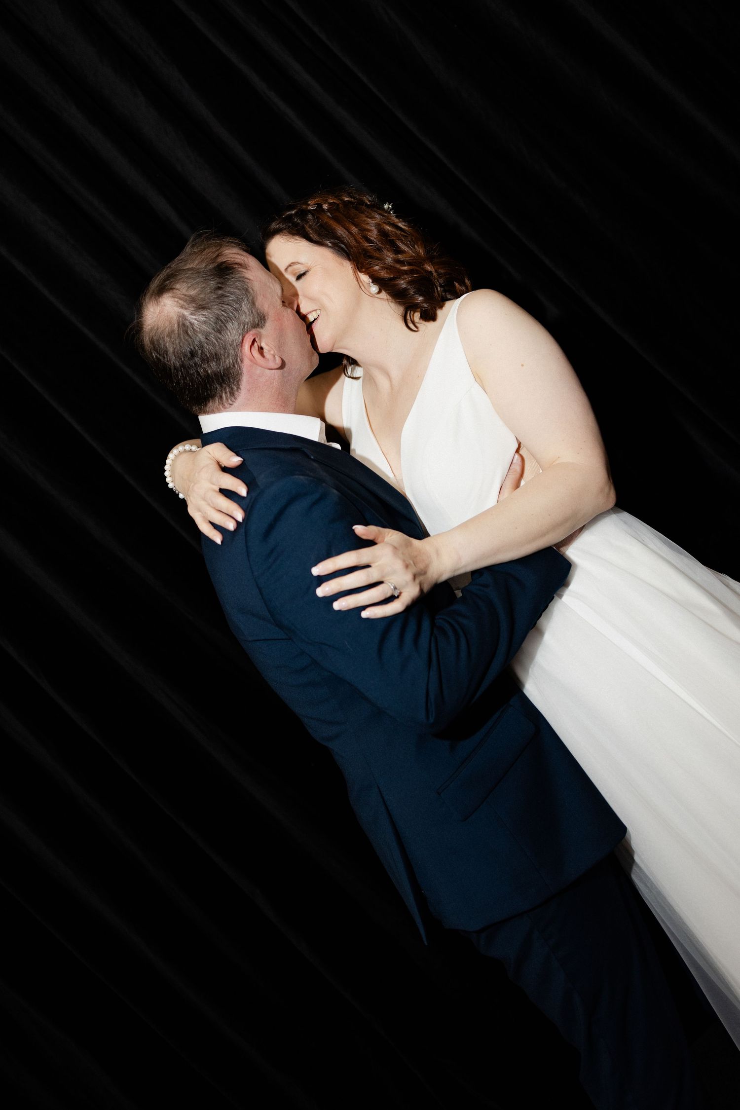A couple in formal wedding attire share an intimate dance pose against a dark background.