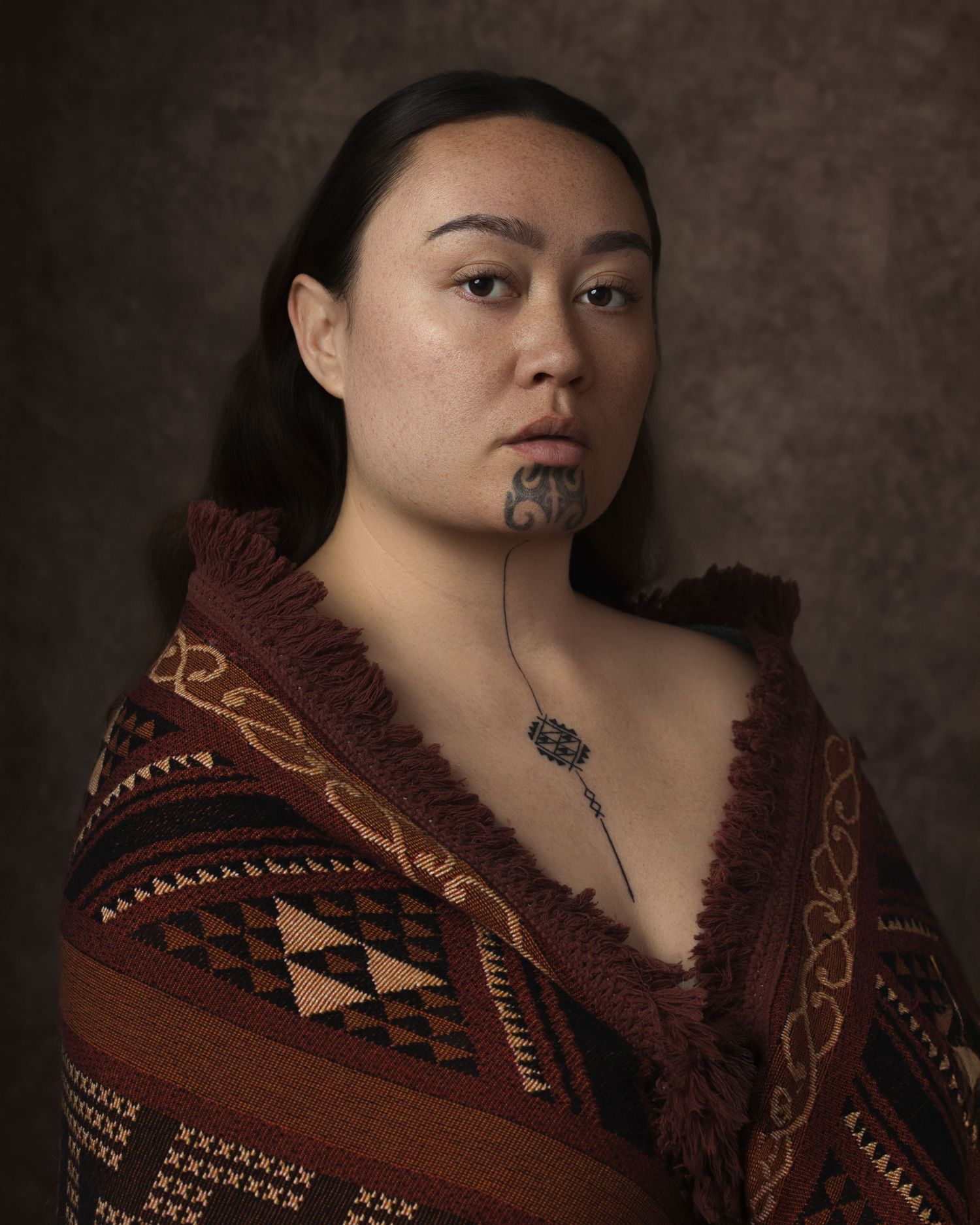 Maori woman wearing traditional korowai cloak and moko kauae chin tattoo, posing for a formal portrait.