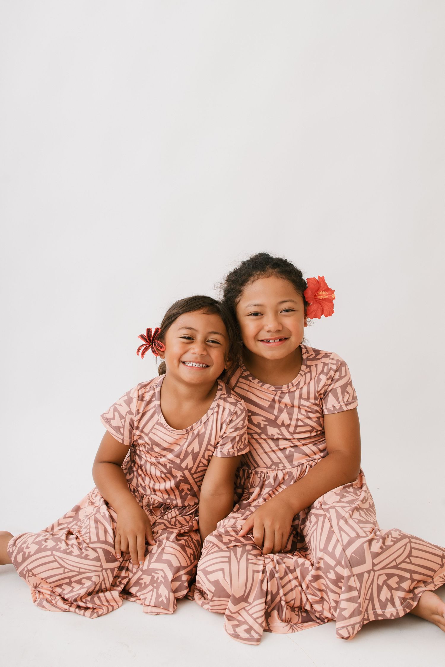 Two children wearing matching pink and white patterned dresses sit together with red flowers in their hair against white background.