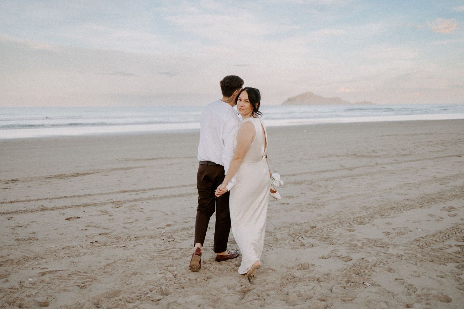 A couple walks together on a scenic beach at sunset wearing wedding attire with mountains in the distance.