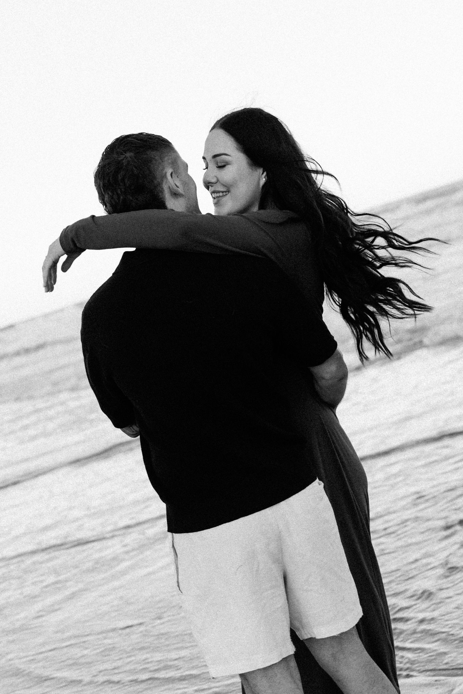 Black and white photo of a couple embracing and smiling on a beach with ocean waves in the background.