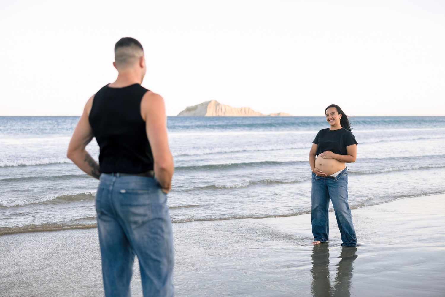 Two people standing on a beach at sunset with waves and mountains in the distance.