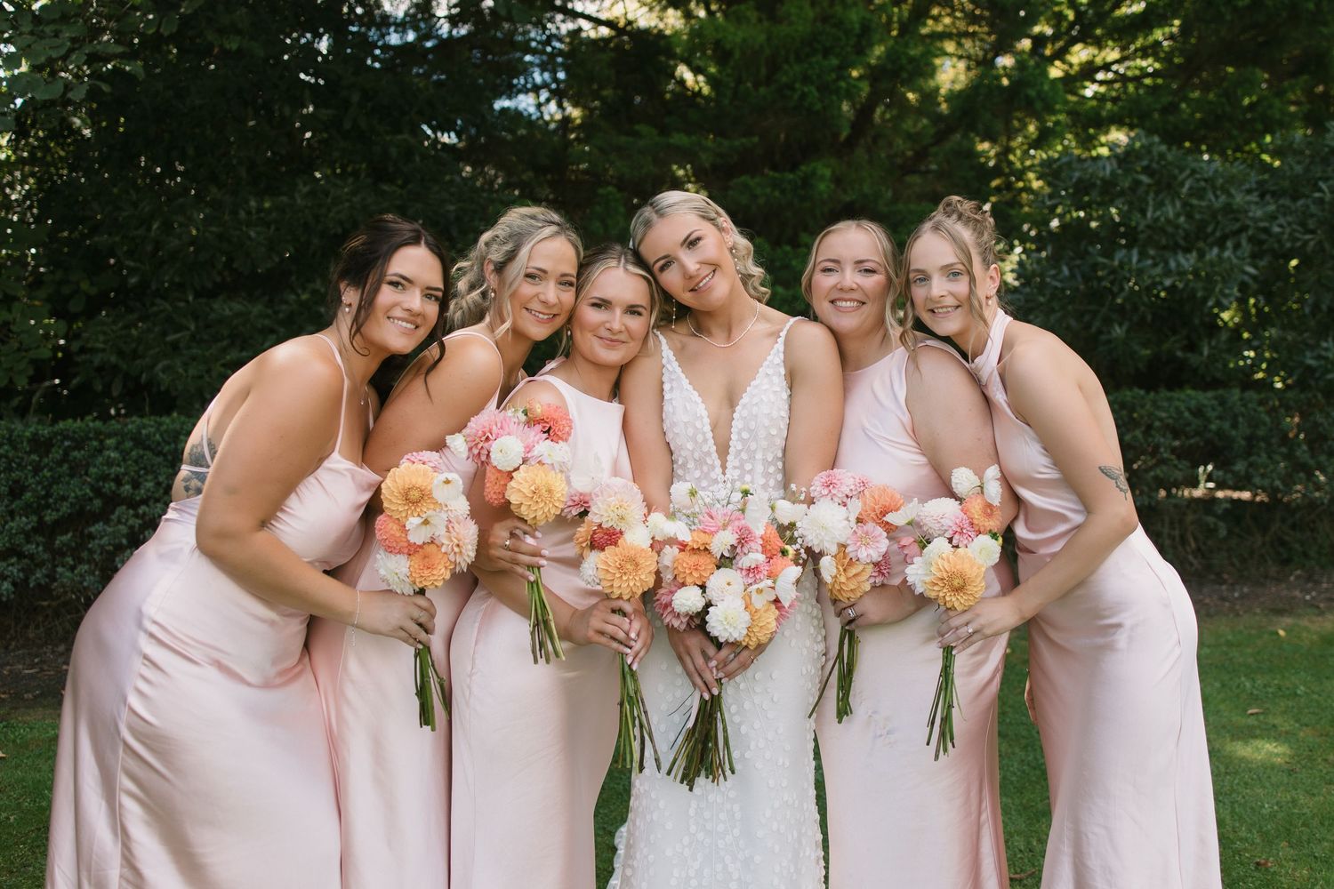 A bridal party in blush pink dresses poses together outdoors holding peach and coral floral bouquets.